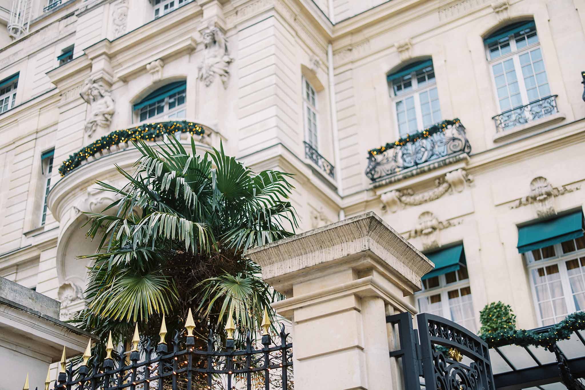 Exterior architectural shot of a grand Haussmann-style Parisian building featuring cream stone facades, ornate sculptural detailing, teal-painted window frames and awnings, and wrought iron balconies with decorative ironwork. The building's balconies and entrance gate are dressed with dark green garland and warm gold fairy lights, suggesting a winter or festive-season event. A large palm tree rises in front of the black and gold-tipped wrought iron entrance gate, with a glazed canopy entrance visible to the right. The composition is an upward-angle wide shot focused on the building's facade and entrance. Potential venue feature image.