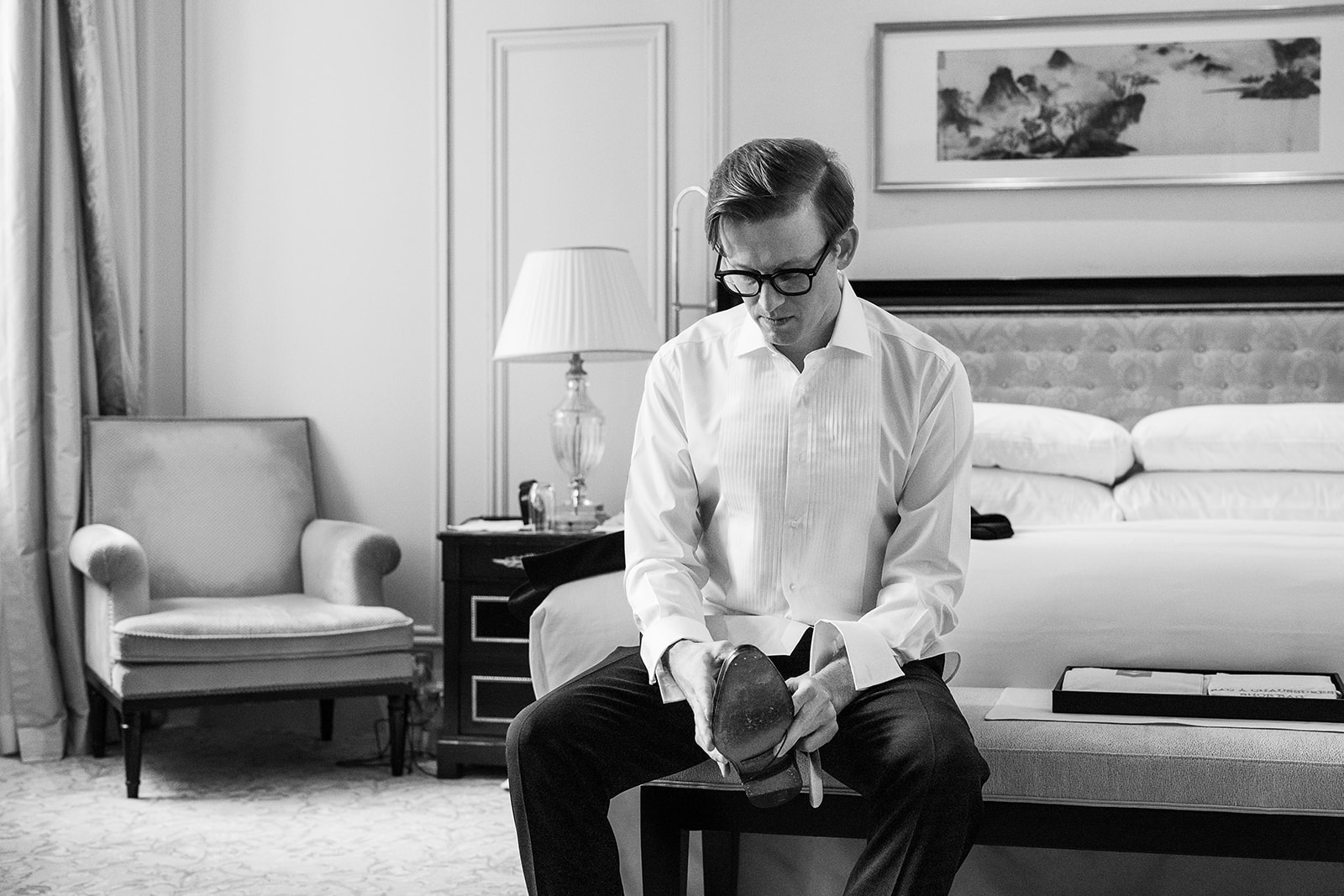 A black-and-white getting-ready shot of the groom seated on a bench at the foot of a hotel bed, putting on his dress shoes. He is wearing a white pleated-front tuxedo shirt with French cuffs and dark formal trousers, and sports black-framed glasses. The room features classic hotel furnishings including an upholstered armchair, a crystal table lamp on a dark bedside cabinet, a patterned headboard, and a framed landscape artwork on the wall. The image is a medium portrait shot with soft, even tones and moderate contrast.