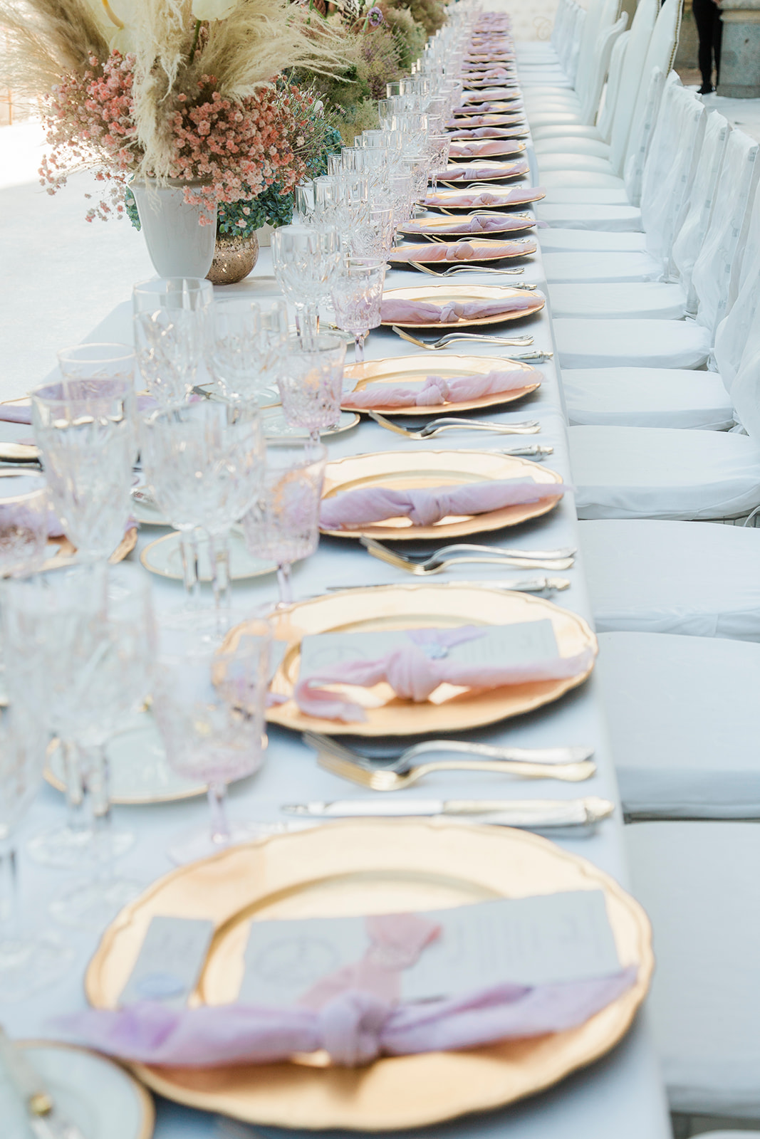 A close-up detail shot of a long reception banquet table set for a large number of guests, appearing to be outdoors or under an open-air structure. The table is dressed with a pale blue-grey linen runner and features gold charger plates topped with dusty rose dinner plates, each place setting styled with a lavender-lilac knotted napkin and a grey menu card. Crystal glassware in both clear and pale lavender-tinted styles is arranged at each setting, alongside silver cutlery. Centerpieces at the far end of the table include arrangements of blush and dusty pink dried and fresh florals — likely dried baby's breath, pampas grass, and small clustered blooms — in white and gold vessels. White chair covers line the right side of the table. The overall decor palette combines dusty blue, gold, blush, and lavender in a soft, romantic classic style.