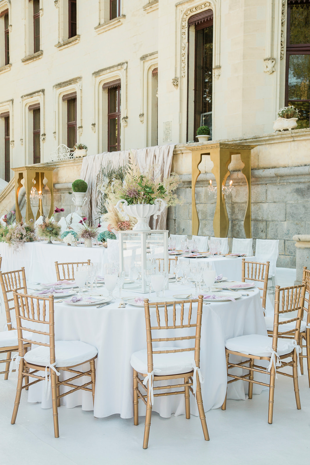 An outdoor wedding reception setup on a terrace or courtyard in front of a classical French chateau with ornate stone facade and tall arched windows. The foreground shows a round guest table dressed in a white linen tablecloth, surrounded by gold chiavari chairs with white cushion pads tied with ribbon, set with clear glassware, gold cutlery, and soft lavender-pink folded napkins on white plates. The centerpiece is a tall white urn vase holding an arrangement of dried and fresh mixed florals including pampas grass, cream astilbe, blush thistle, and greenery, alongside a clear glass hurricane lantern. In the background, a long head table is visible with similar decor including smaller low floral arrangements in muted mauve, dusty blue, and cream tones, flanked by gold ornate decorative pedestals with lit candelabras and a draped blush-taupe fabric backdrop panel. The overall palette is white, gold, and soft pastels with a classic, romantic styling approach. Wide shot with a slightly elevated angle. Potential venue feature image.