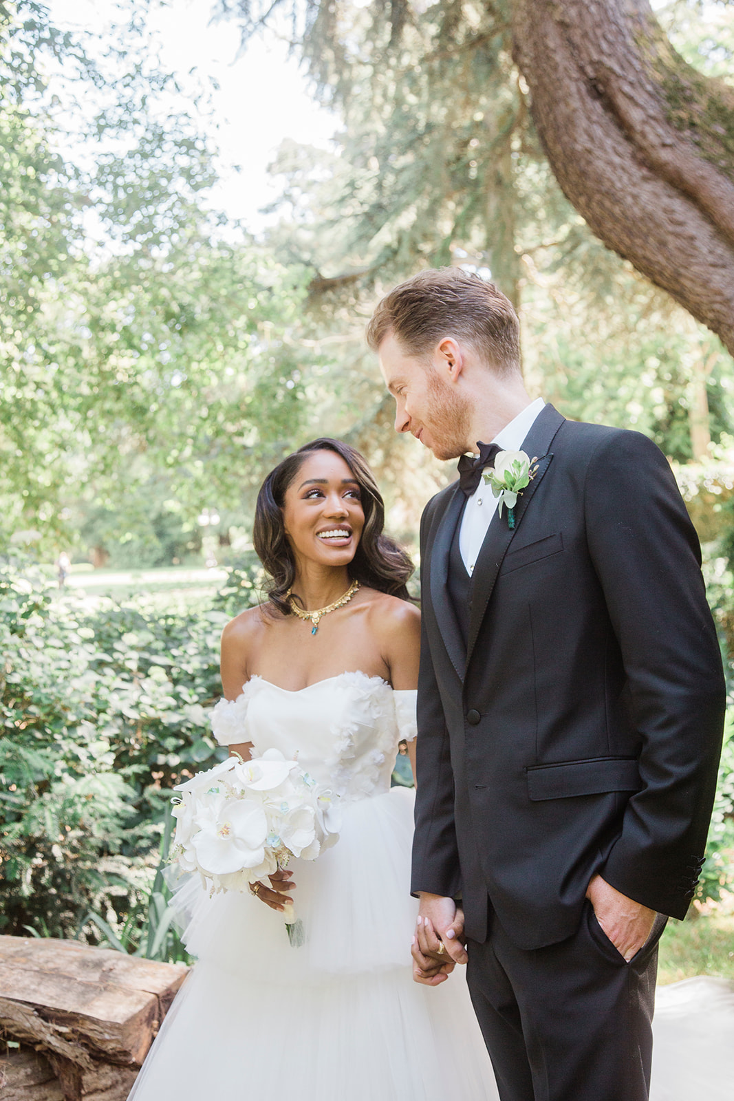 Outdoor couple portrait of a bride and groom holding hands and facing each other along a garden path. The bride wears an off-the-shoulder white ballgown with textured floral appliqué detailing on the bodice, paired with a gold and blue pendant necklace; she carries a cascading bouquet of white phalaenopsis orchids. The groom wears a black tuxedo with satin lapels, a black bow tie, and a boutonnière of white blooms with small green succulents. The setting is a lush garden with mature trees and dense greenery in soft natural daylight. The shot is a mid-length portrait with a slightly bright, airy exposure.