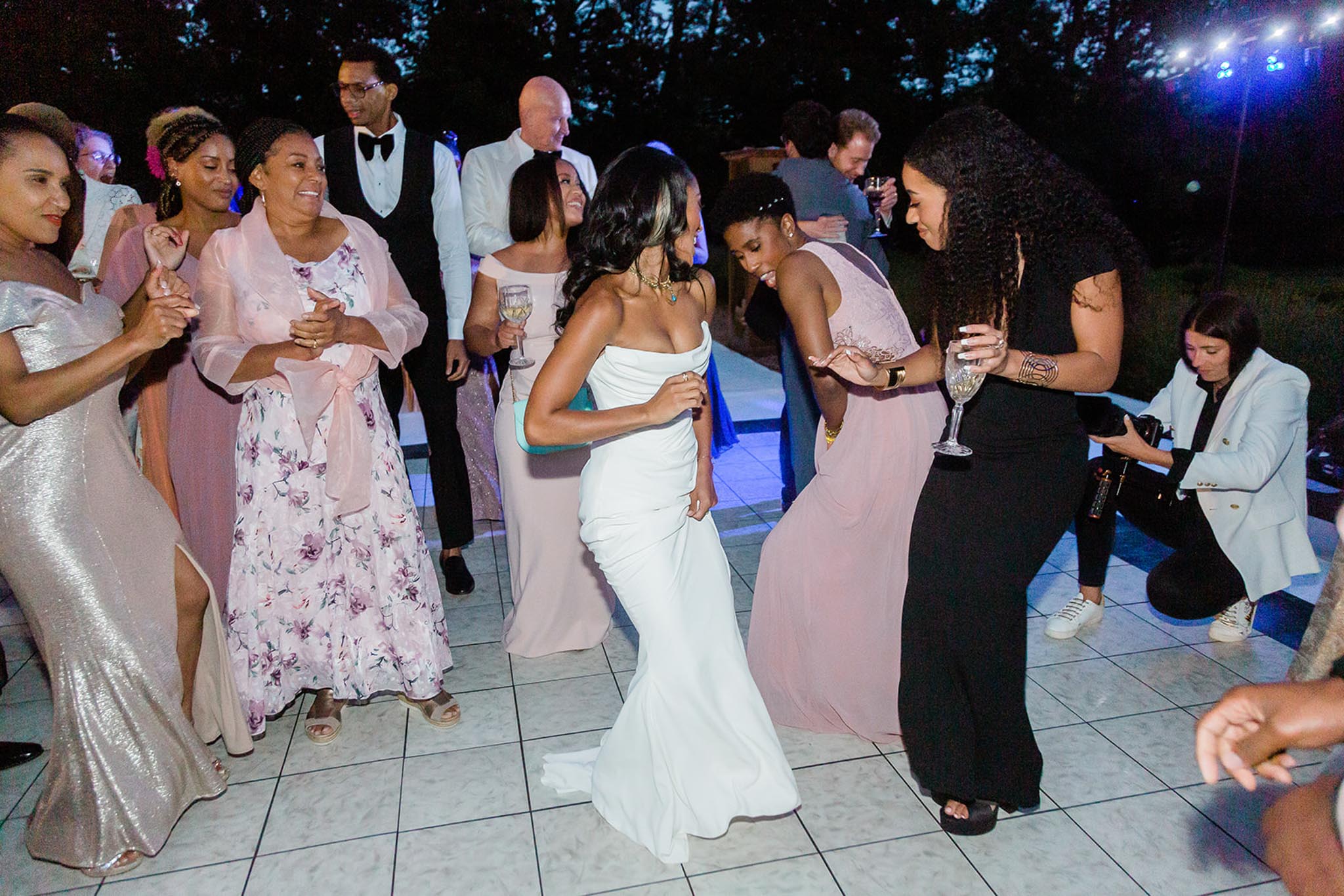 An outdoor evening reception dance floor scene featuring the bride dancing energetically among approximately 10–12 guests. The bride wears a fitted off-the-shoulder white gown with a sleek silhouette and a small train. She is surrounded by guests in varied attire including a blush pink chiffon floor-length bridesmaid dress, a silver sequined gown, a floral maxi dress in lavender and blush, and a black sleeveless dress. The dance floor is a white tiled portable floor lit with blue uplighting, set outdoors at night with a dark tree line visible in the background. A photographer crouches at the edge of the frame capturing the moment. The overall styling is modern and polished, and the atmosphere is lively with guests laughing and holding champagne glasses. Wide candid shot taken at eye level.