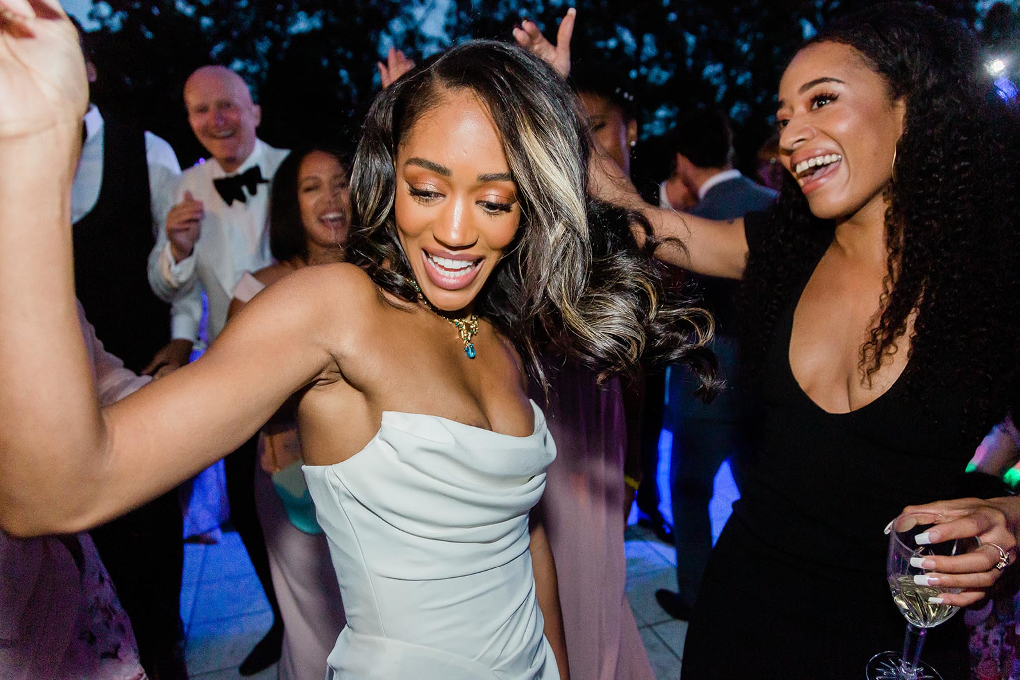 An outdoor evening wedding reception dance floor scene with a large group of guests dancing together. The bride is centered in the foreground, wearing a strapless ivory structured satin gown with ruched bodice and a gold and blue gemstone pendant necklace, her highlighted dark hair in loose waves. She is dancing energetically and smiling. To her right, a female guest in a black deep-V dress holds a wine glass and laughs. Behind them, additional guests are visible including a man in a black tuxedo with bow tie and a woman in a floral dress. Blue-toned event lighting illuminates the dance floor, giving the background a vibrant nighttime party atmosphere. The composition is a close-up candid portrait shot capturing the energy of the reception.