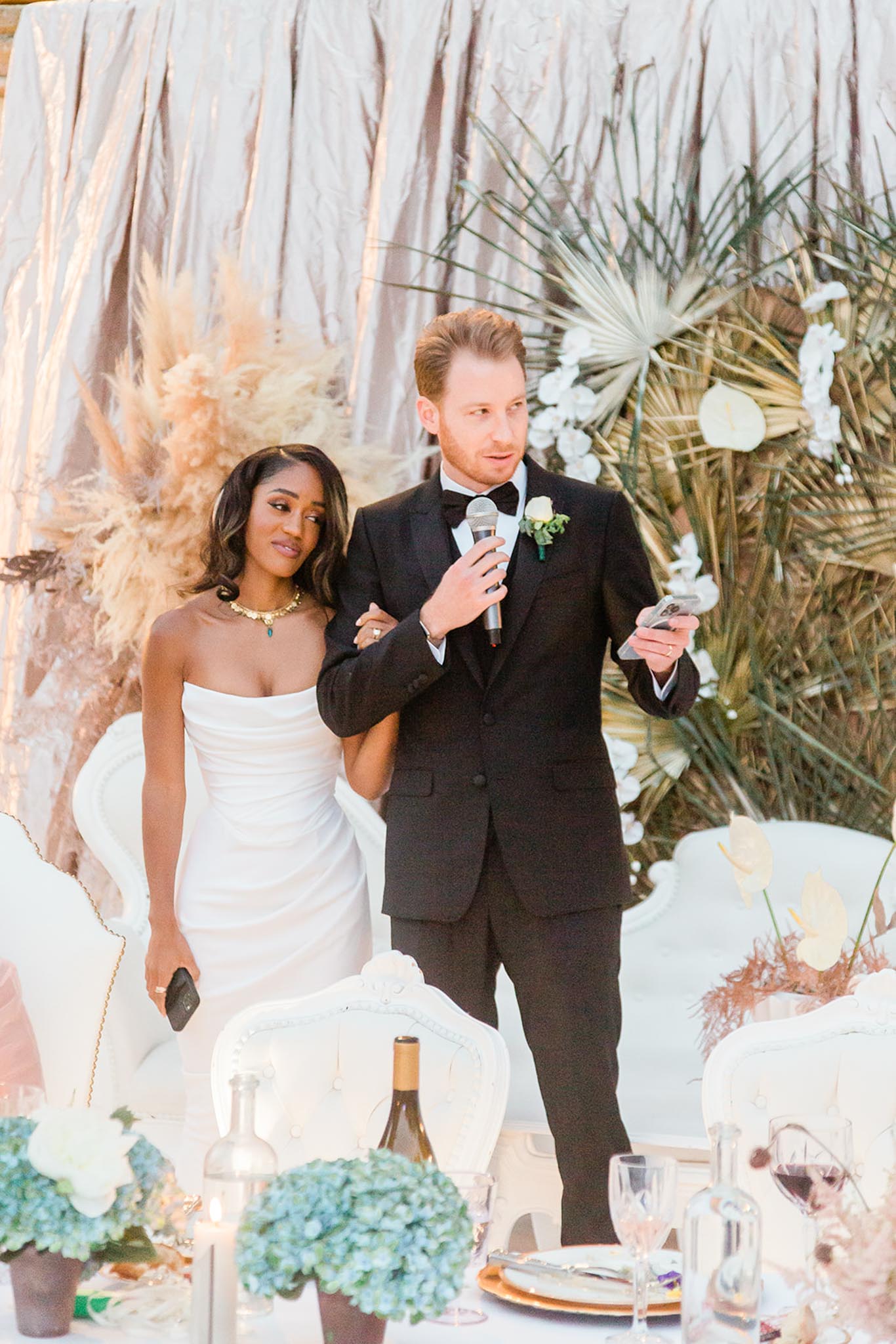 The couple stands together at what appears to be an outdoor reception, with the groom holding a microphone and a phone, suggesting he is giving a speech. The bride wears a strapless white fitted gown and a gold statement necklace with a green stone pendant, while the groom is dressed in a black tuxedo with a bow tie and a white boutonniere accented with greenery. Behind them is a decorative backdrop combining ivory draped fabric, pampas grass, dried palm fronds, and white orchids, creating a boho-modern aesthetic in a neutral and earth-tone palette. The foreground shows a sweetheart table set with white upholstered French-style chairs, blue hydrangea arrangements, crystal glassware, gold-rimmed charger plates, and candles; the shot is a medium portrait-style image capturing both the couple and the styled table decor.