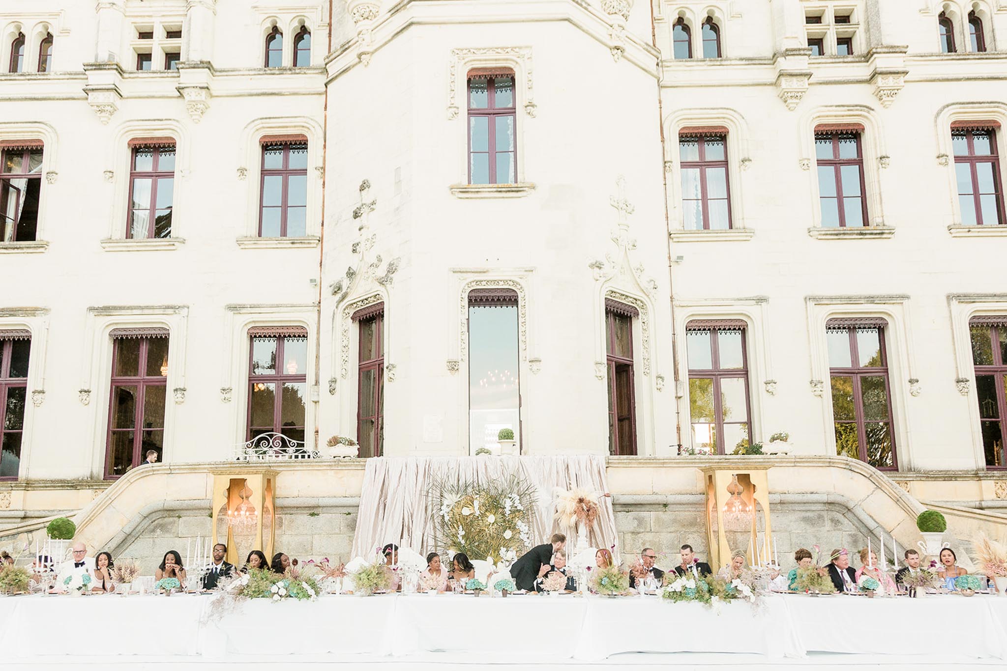 An outdoor wedding reception dinner is set up directly in front of a large French chateau with cream-colored stone facade, ornate architectural detailing, and burgundy-framed windows and doors. A long head table seats approximately 25–30 guests, with the couple positioned at the center where the groom leans toward the bride; the table is draped in blush pink linen and backed by a voluminous blush fabric installation flanked by tall gold-framed decorative panels with amber-toned lanterns. The tablescape features clusters of blue-green hydrangeas, blush and ivory florals, pampas grass, dried botanicals, tall white taper candles, and a large circular gold floral arrangement as the central backdrop piece. The shot is a wide, slightly elevated perspective that frames the full facade of the chateau above the reception scene. Potential venue feature image.