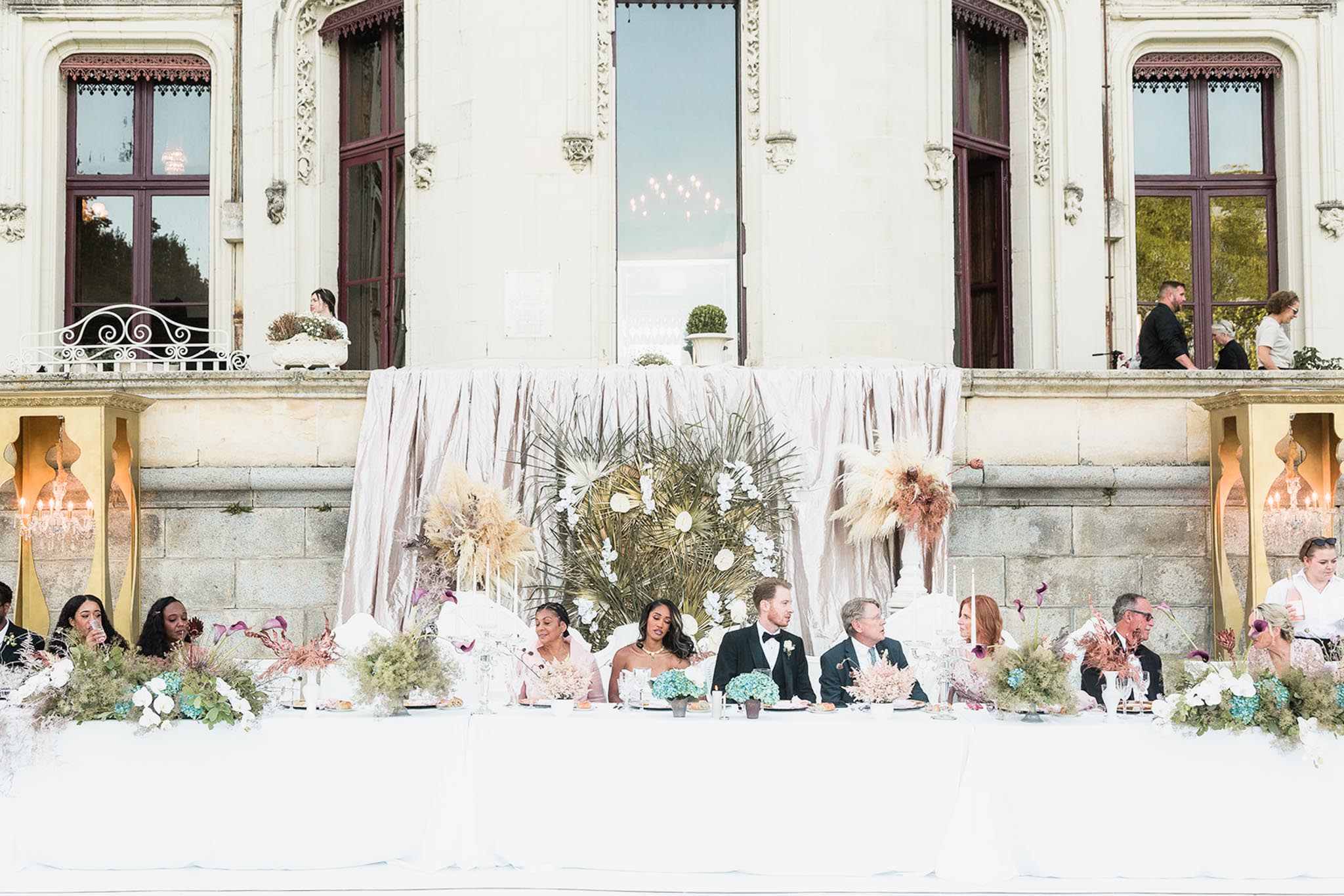 An outdoor wedding reception head table is set directly against the ornate stone facade of a French chateau, with approximately ten guests and the couple seated along a white linen-draped long table. The bride wears a strapless white gown and the groom wears a black tuxedo with bow tie, seated at center; a bridesmaid in blush pink sits to the bride's left. The backdrop features draped blush-toned fabric panels flanking a large sculptural floral installation composed of dried palm leaves, pampas grass, white orchids, and gold accents. Table centerpieces include a mix of teal blue hydrangeas, dried blush and terracotta florals, white orchids, and loose greenery, with tall white taper candles; gold candelabras with lit candles anchor each end of the table. The overall decor palette blends blush, teal, terracotta, and gold in a modern botanical style, and the wide shot captures the full table symmetrically framed by the chateau's arched windows and balcony above. Potential venue feature image.