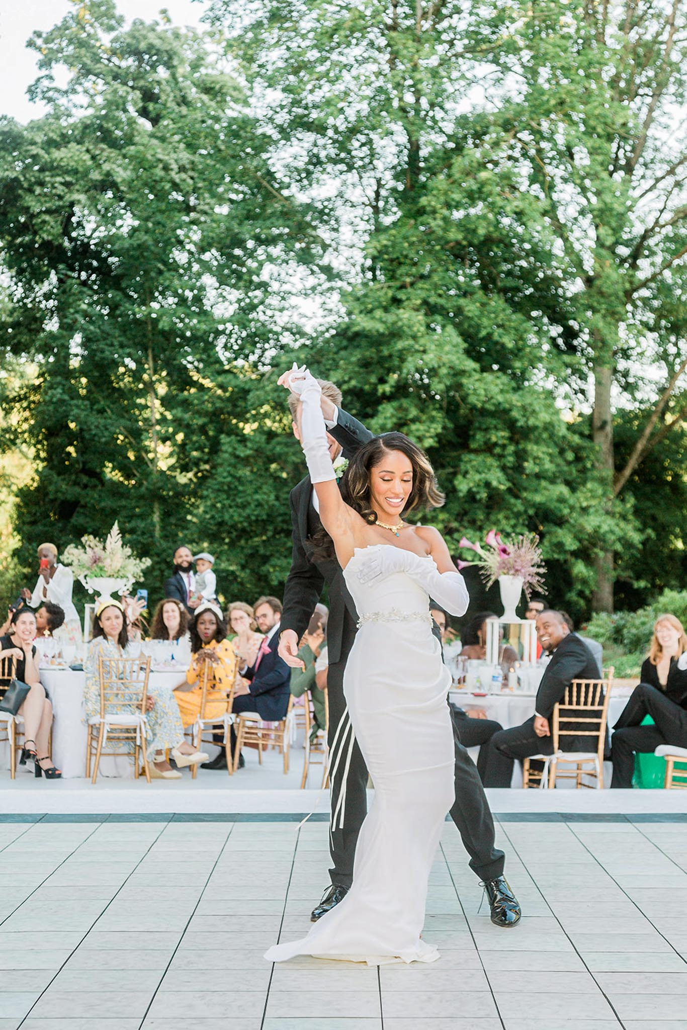 The couple is performing their first dance on an outdoor pale grey dance floor during a reception, with the groom dipping and spinning the bride as she smiles broadly. The bride wears a fitted strapless white gown with a floral detail at the bust, a crystal-embellished waist belt, and long white satin gloves, accessorized with a yellow gemstone necklace; the groom is dressed in a black tuxedo. Approximately 20–25 guests are seated at round tables in the background on gold chiavari chairs, watching and reacting to the dance, with tall white pedestal floral arrangements featuring pampas grass and pink blooms visible between tables. The shot is a medium full-length portrait taken at ground level, capturing the energy of the dance moment during an outdoor evening garden reception.