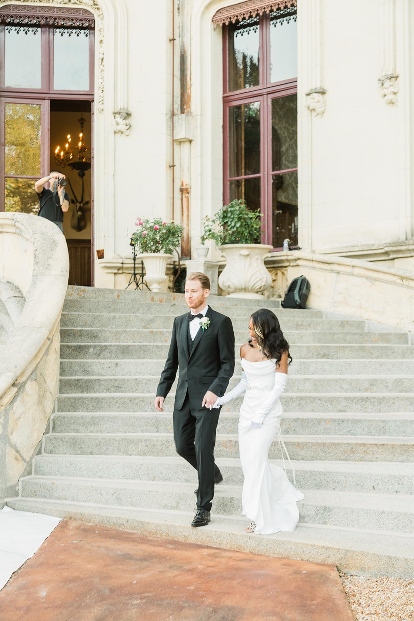 A couple walks hand-in-hand down the grand stone staircase of a French chateau, captured in a full-length portrait shot. The groom wears a black tuxedo with a black bow tie and a small white and green boutonniere, while the bride wears a fitted off-the-shoulder white gown with long white opera gloves and a small train. The chateau facade features ornate cream stonework, tall burgundy-framed windows, and large stone urns planted with pink roses and greenery; a chandelier is visible through the open doorway. A photographer is visible in the background on the steps, photographing the couple from a different angle, and lighting equipment is also partially visible. The styling is classic and modern, with a black-and-white color palette for the couple's attire.
