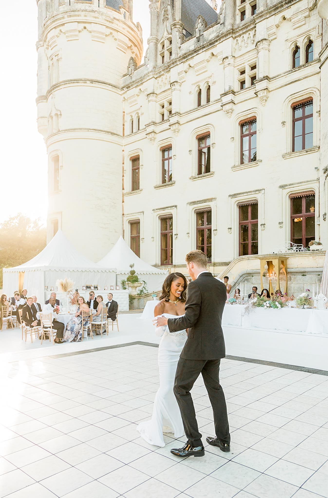 The bride and groom share their first dance on an outdoor tiled dance floor in front of a grand French chateau with a round tower, ornate stone facade, and dark red-framed windows bathed in warm golden-hour light. The bride wears a white strapless fitted gown with a beaded waistband and a gold statement necklace, while the groom is dressed in a dark suit with black dress shoes. Approximately 20–30 guests are seated at gold chiavari chairs around reception tables decorated with colorful floral centerpieces and tall white taper candles, with a white marquee tent serving as a bar area in the background. The wide-angle shot captures both the couple dancing in the foreground and the full outdoor reception setting against the chateau exterior. Potential venue feature image.