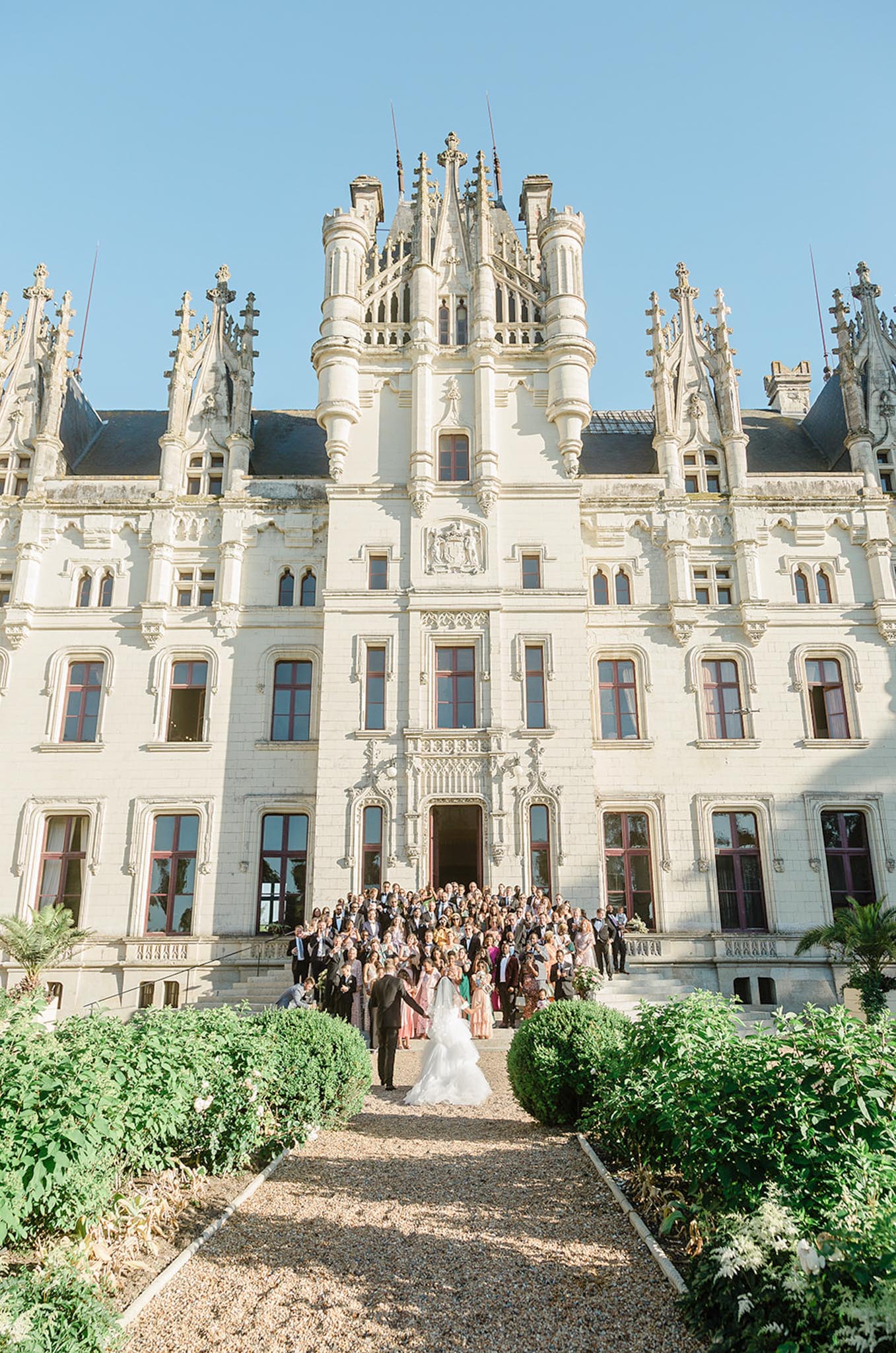 A wide-angle group portrait taken outdoors in front of a large French Gothic-style chateau with white stone facades, ornate spires, turrets, and red-framed windows. The couple stands in the foreground on a gravel path — the bride in a full white ballgown with a long veil and the groom in a dark suit — holding hands with their backs partially toward the camera as they face approximately 100–150 guests arranged on the chateau's front steps. Guests are dressed in a mix of formal attire including colorful dresses in blush, coral, and green tones alongside dark suits and tuxedos. The composition is a full vertical wide shot that emphasizes the scale of the chateau architecture above the assembled wedding party. Potential venue feature image.
