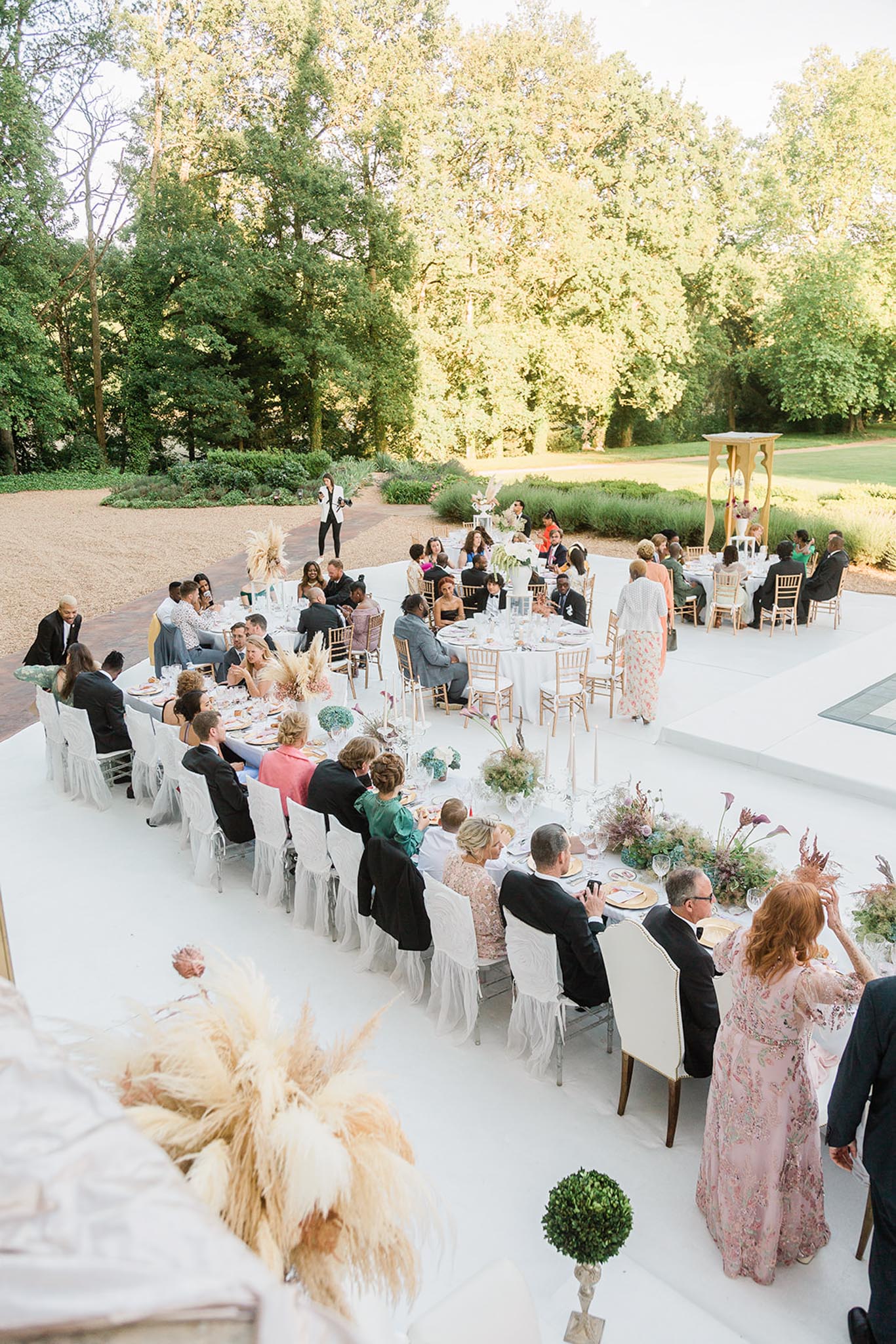 An outdoor wedding reception dinner is underway on a white-painted terrace or courtyard, with approximately 60–70 guests seated across a mix of long rectangular tables and round tables arranged in a loose horseshoe configuration. The long head table is dressed in white linens with white tulle-skirted chairs and ivory upholstered armchairs, decorated with a lush runner of dried pampas grass, burgundy calla lilies, dusty pink proteas, sage greenery, and muted mauve florals. Round tables in the background feature chiavari chairs and white linens with similar arrangements including tall pampas grass centerpieces. Guests are dressed in formal and semi-formal attire including black tuxedos, a blush floral gown, emerald dress, coral outfit, and various colorful evening wear. A person in a white blazer stands near the back, appearing to address the group, suggesting a speech or performance moment. Boxwood topiaries flank the foreground, and a decorative gold standing structure is visible to the right. The overall decor palette is neutral and warm with dusty rose, ivory, sage, and dried botanicals in a modern romantic style. Wide overhead shot taken from an elevated angle.