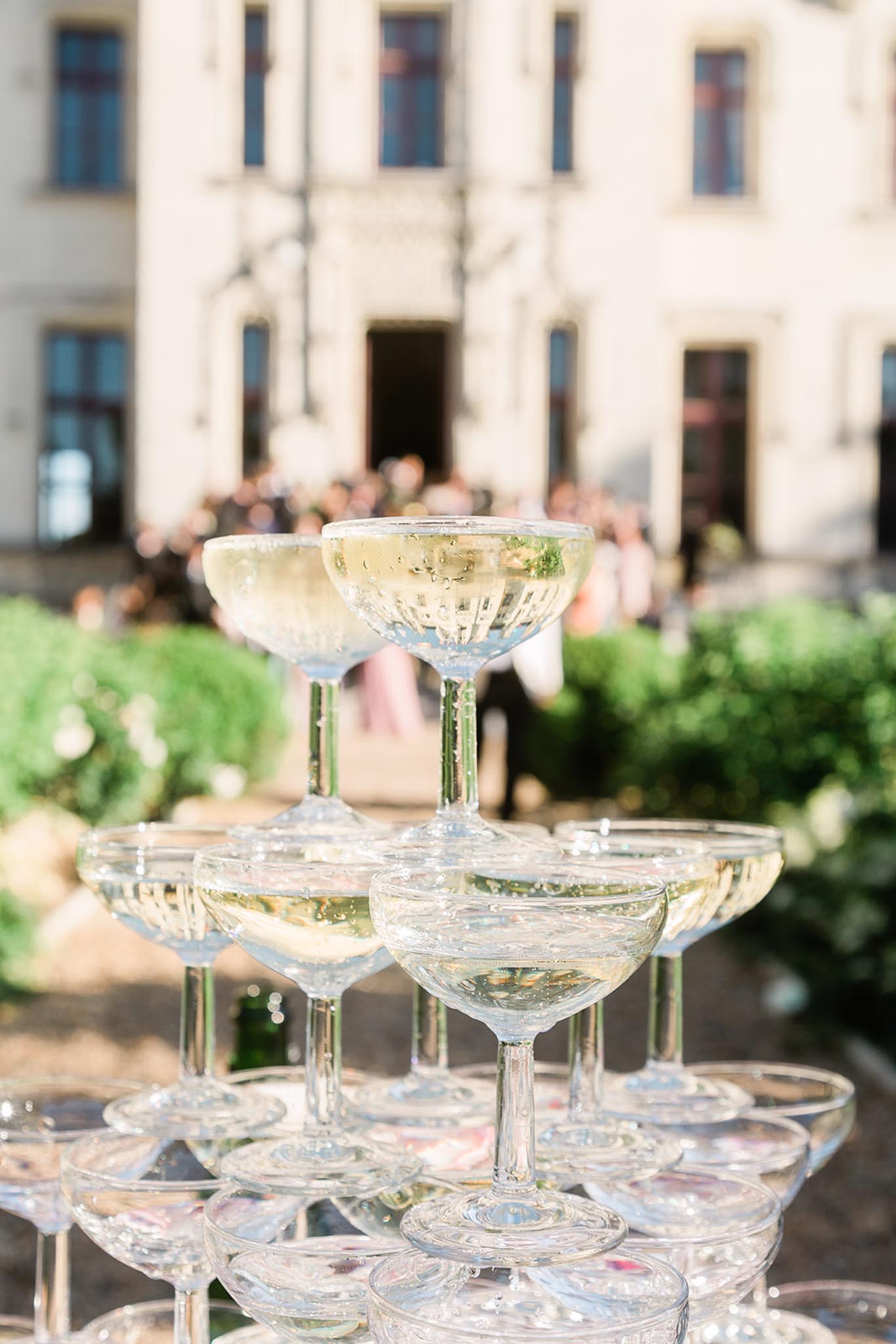A close-up detail shot of a champagne tower made from classic coupe glasses filled with pale golden champagne, set up outdoors during a cocktail hour. The tower consists of at least three visible tiers, with the glasses stacked in the traditional pyramid formation. In the soft-focus background, a large pale stone château-style building is visible with guests gathered near its entrance. The classic coupe glass style and outdoor château setting give the scene a traditional French wedding aesthetic.