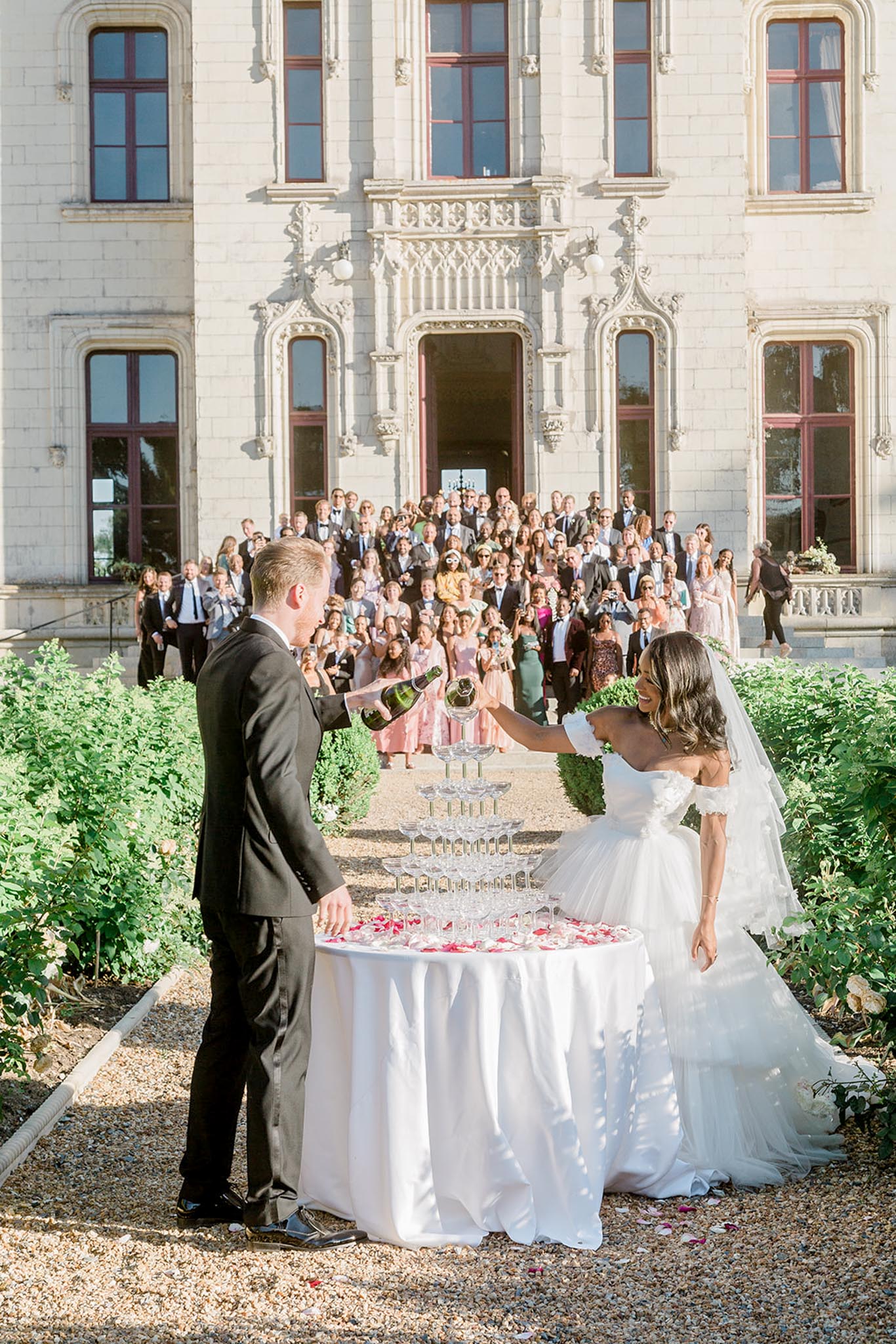 The bride and groom are pouring champagne into a coupe tower during an outdoor cocktail hour in front of a French chateau with ornate limestone Gothic-style architecture and burgundy-trimmed windows. The groom wears a dark charcoal suit, while the bride wears a strapless layered white tulle ballgown with ruffled off-the-shoulder detailing and a long veil, her dark hair worn down in waves. The champagne tower is displayed on a round table with a white linen, scattered with pink and red rose petals at the base. A large crowd of approximately 80-100 guests in formal attire is gathered on the chateau's front steps behind the couple, watching and celebrating. The wide-angle shot captures the full scene from the couple in the foreground to the assembled guests and chateau facade in the background. Potential venue feature image.