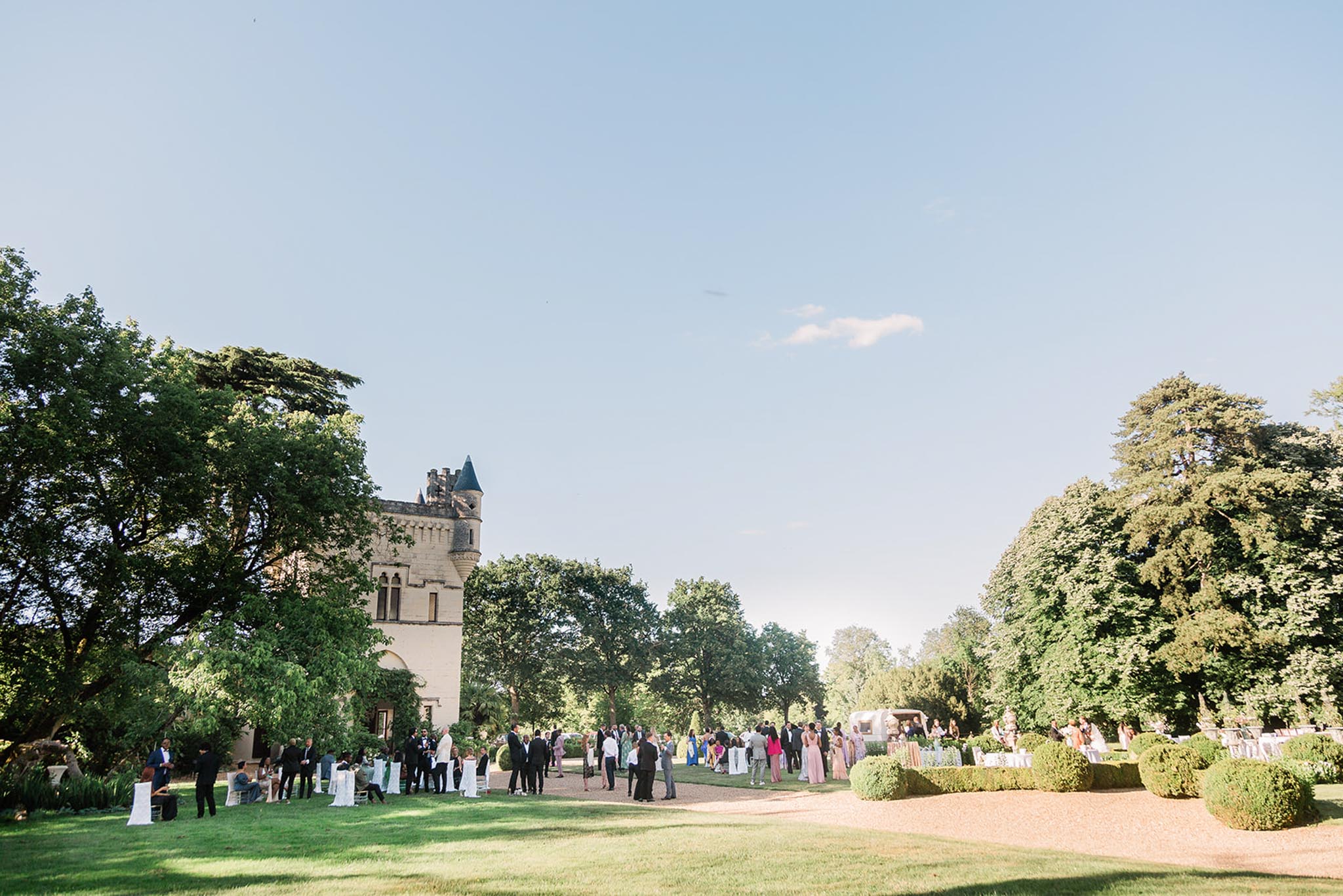 A cocktail hour taking place outdoors on the grounds of a French chateau, with a stone tower featuring a dark conical turret visible on the left side of the frame. Approximately 60–80 guests are mingling across a gravel pathway and lawn area, dressed in formal attire including dark suits and colorful gowns in blush, green, and mixed hues. White chair covers and cocktail-height tables dressed in white linens are visible near the chateau facade, along with additional seating and decor arrangements further along the path. The image is a wide environmental shot taken in bright daytime light, capturing the full scale of the venue grounds with manicured box hedges lining the gravel path. Potential venue feature image.