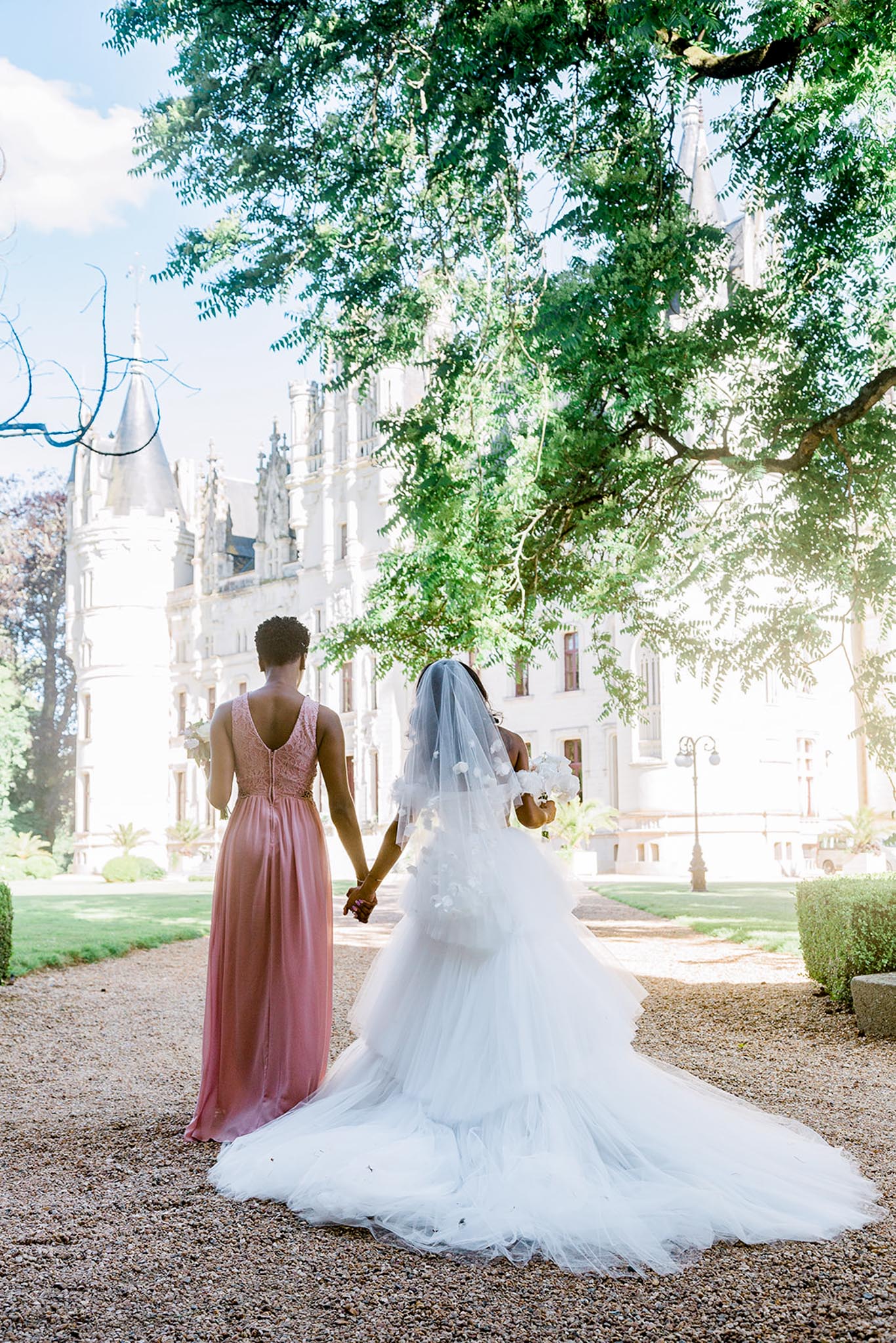 A bride and bridesmaid are photographed from behind, holding hands and facing a white French château with Gothic-style turrets and ornate stone facade. The shot is taken outdoors on a gravel pathway in the château grounds. The bride wears a full white tulle ballgown with a cathedral-length train and a veil adorned with floral appliqués, and carries a white bouquet. The bridesmaid wears a floor-length dusty rose gown with a lace V-back bodice and chiffon skirt. The composition is a wide portrait shot with the château prominently centered in the background. Potential venue feature image.