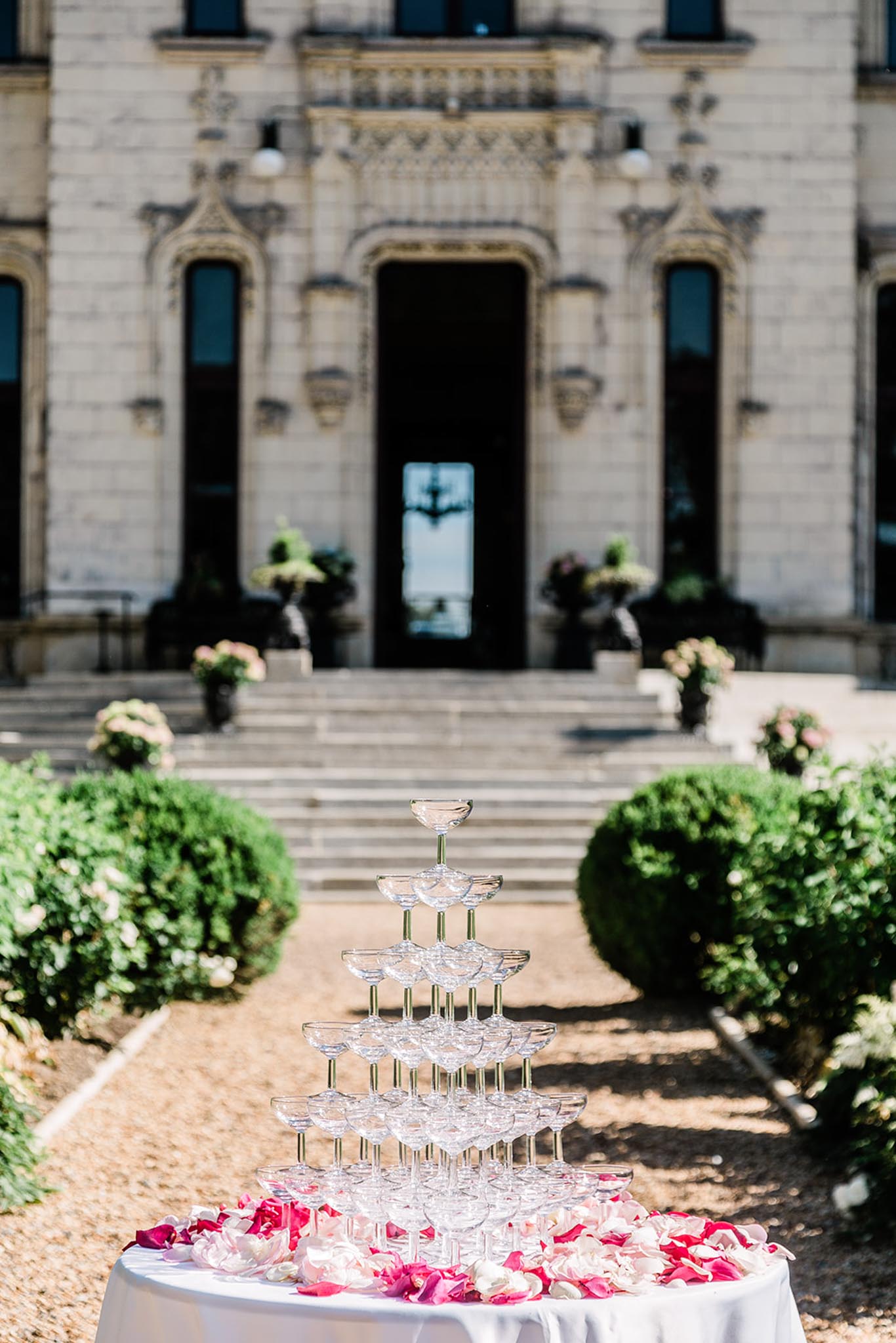 A champagne tower composed of multiple tiers of coupe glasses is displayed outdoors on a white linen-covered cocktail table, surrounded by scattered deep fuchsia and blush pink rose petals. The setup is positioned in front of a grand French chateau with ornate carved stone facade, wide entry steps, potted topiaries, and black decorative urns with greenery flanking the entrance. The foreground is in sharp focus while the chateau facade is softly blurred in the background, creating depth. The composition is a medium portrait shot taken in natural daylight. Potential venue feature image.