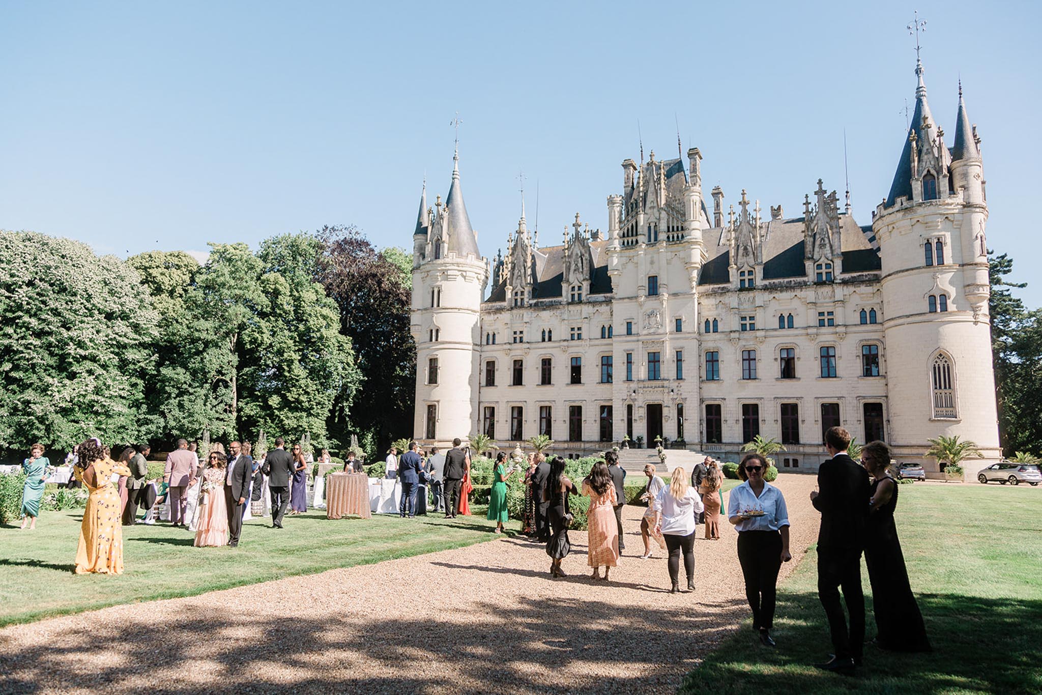 A wedding cocktail hour taking place on the grounds of a large French Gothic-style chateau, featuring cream-colored stone facades, pointed turrets, ornate dormers, and dark slate roofing. Approximately 30–40 guests mingle outdoors on a gravel path and lawn in front of the chateau, dressed in colorful formal attire including a yellow floral gown, a blush pink dress, a green dress, and various dark suits. High-top cocktail tables with blush or champagne linens are visible among the guests. The shot is a wide-angle view taken from ground level, capturing both the crowd and the full facade of the chateau. Potential venue feature image.