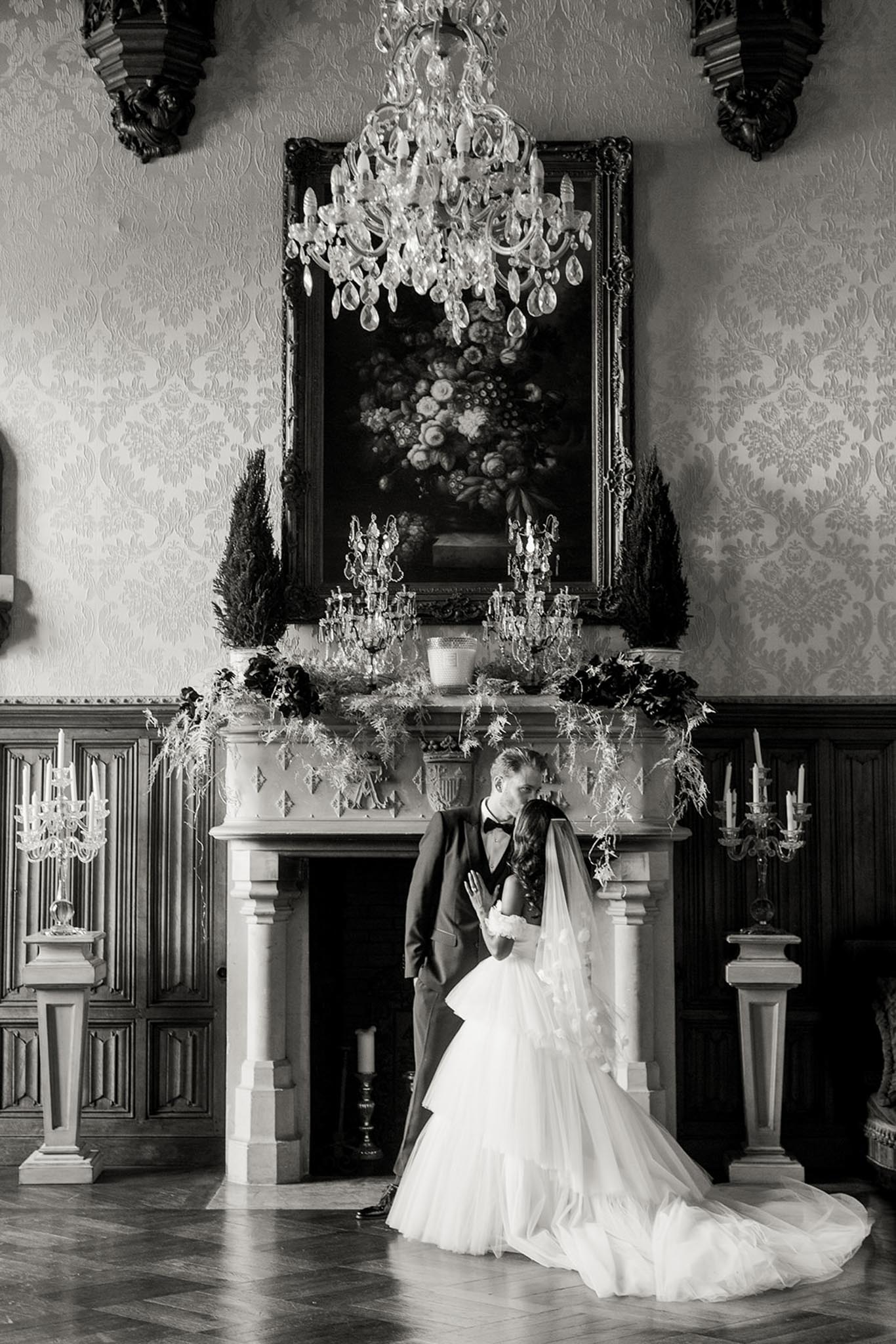 This is a black-and-white portrait of a bride and groom sharing a kiss in front of an ornate stone fireplace inside a formal chateau ballroom or salon. The groom wears a dark tuxedo with a bow tie, and the bride wears a tiered, full-skirted ball gown with a long cathedral veil that trails across the herringbone parquet floor. The fireplace mantel is dressed with a lush garland of dark foliage and fern-like botanicals, flanked by crystal candelabras and tall topiary trees, with a large gilt-framed oil painting of flowers hanging above it. A crystal chandelier hangs overhead, and the walls feature heavily textured damask wallpaper with dark wood wainscoting, creating a classic, formal French interior aesthetic. The image is a full-length portrait shot with strong contrast between the dark architectural elements and the bright whites of the bride's gown and veil. Potential venue feature image.
