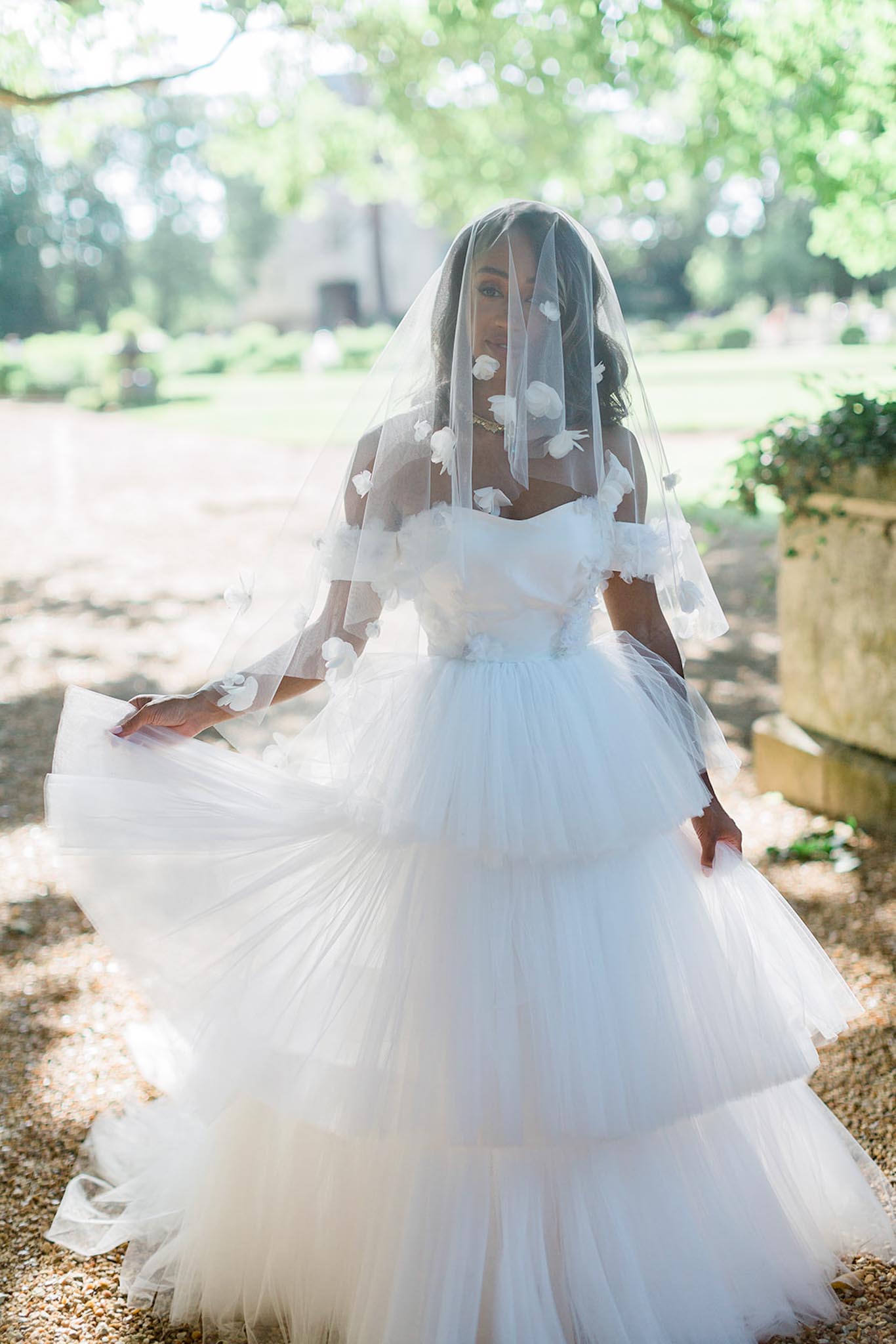 A bridal portrait taken outdoors in a garden setting, with a gravel path and stone planter visible in the background alongside a partially visible stone building. The bride wears a voluminous white tulle ball gown with a tiered, layered skirt and an off-the-shoulder neckline adorned with three-dimensional white floral appliqués. Her veil, which falls over her face, is also embellished with matching white 3D floral details and is caught in motion, partially swept to one side. The shot is a mid-length portrait with the dress skirt filling much of the lower frame, captured in a slightly editorial, movement-driven style.