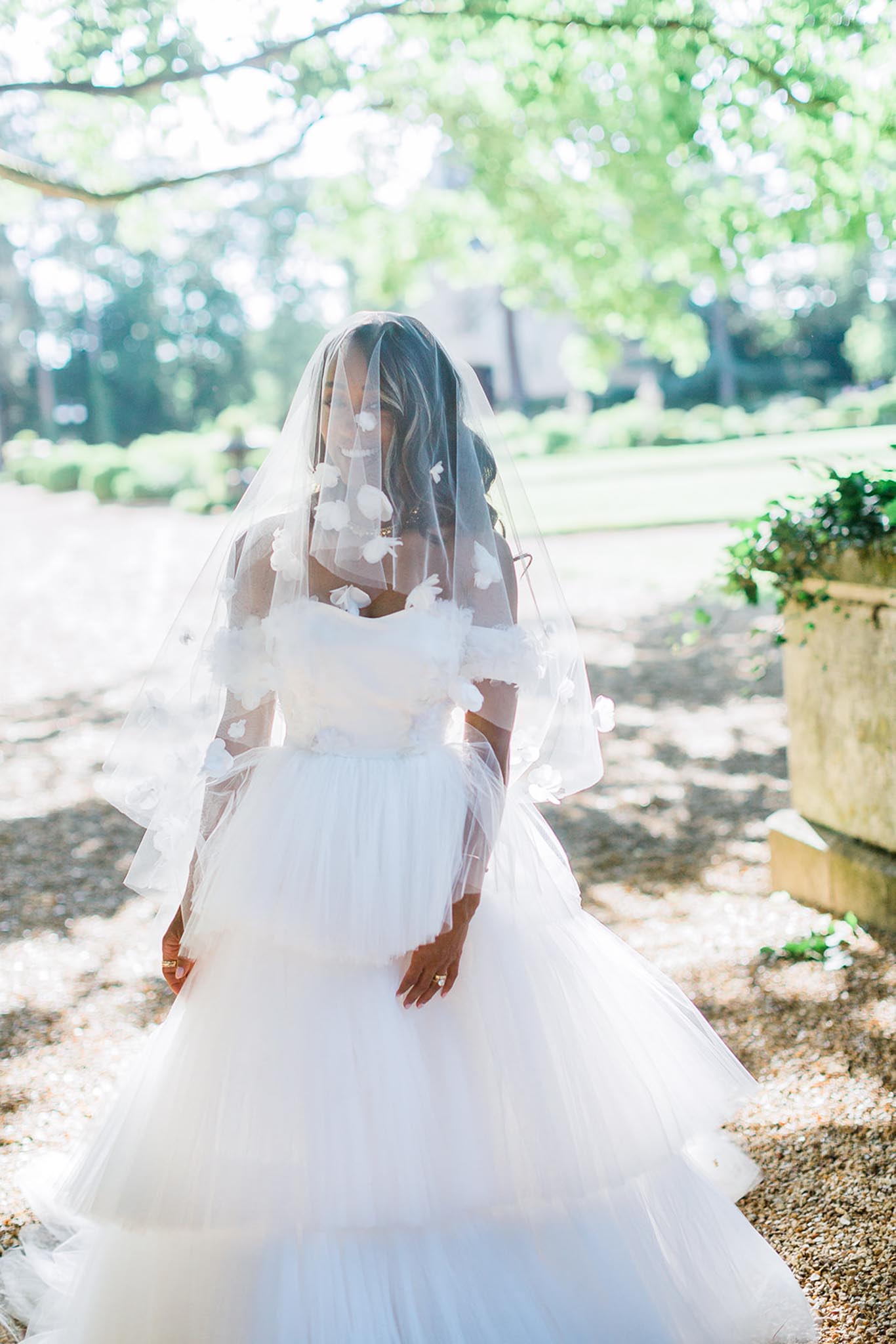 A bridal portrait taken outdoors in a formal garden setting, with a gravel path, trimmed hedges, and a stone urn visible in the background. The bride wears a voluminous white tulle ball gown with 3D floral appliqués along the off-the-shoulder neckline and bodice, paired with a cathedral-length veil adorned with scattered white floral or butterfly appliqués draped over her face. She stands facing the camera with her hands clasped in front of her, wearing a gold ring and an engagement ring. The image is a mid-length portrait shot with soft, dappled natural light filtering through the tree canopy above.