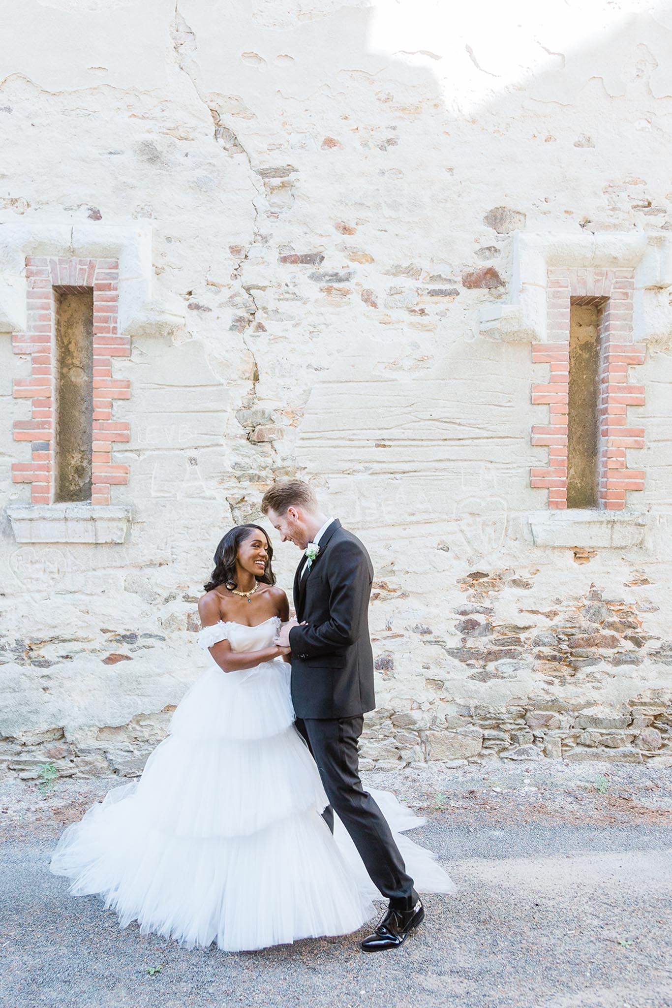 A couple portrait shot outdoors against a weathered stone and brick wall with exposed plasterwork and recessed window niches. The bride wears an off-the-shoulder white tulle ballgown with tiered ruffled skirt and a gold statement necklace, while the groom is dressed in a dark charcoal suit with a white dress shirt, bow tie, and a small white boutonniere. The two are facing each other closely, smiling and laughing, with the groom leaning slightly toward the bride. The composition is a full-length portrait in natural bright daylight, capturing the full volume of the gown against the rustic architectural backdrop.