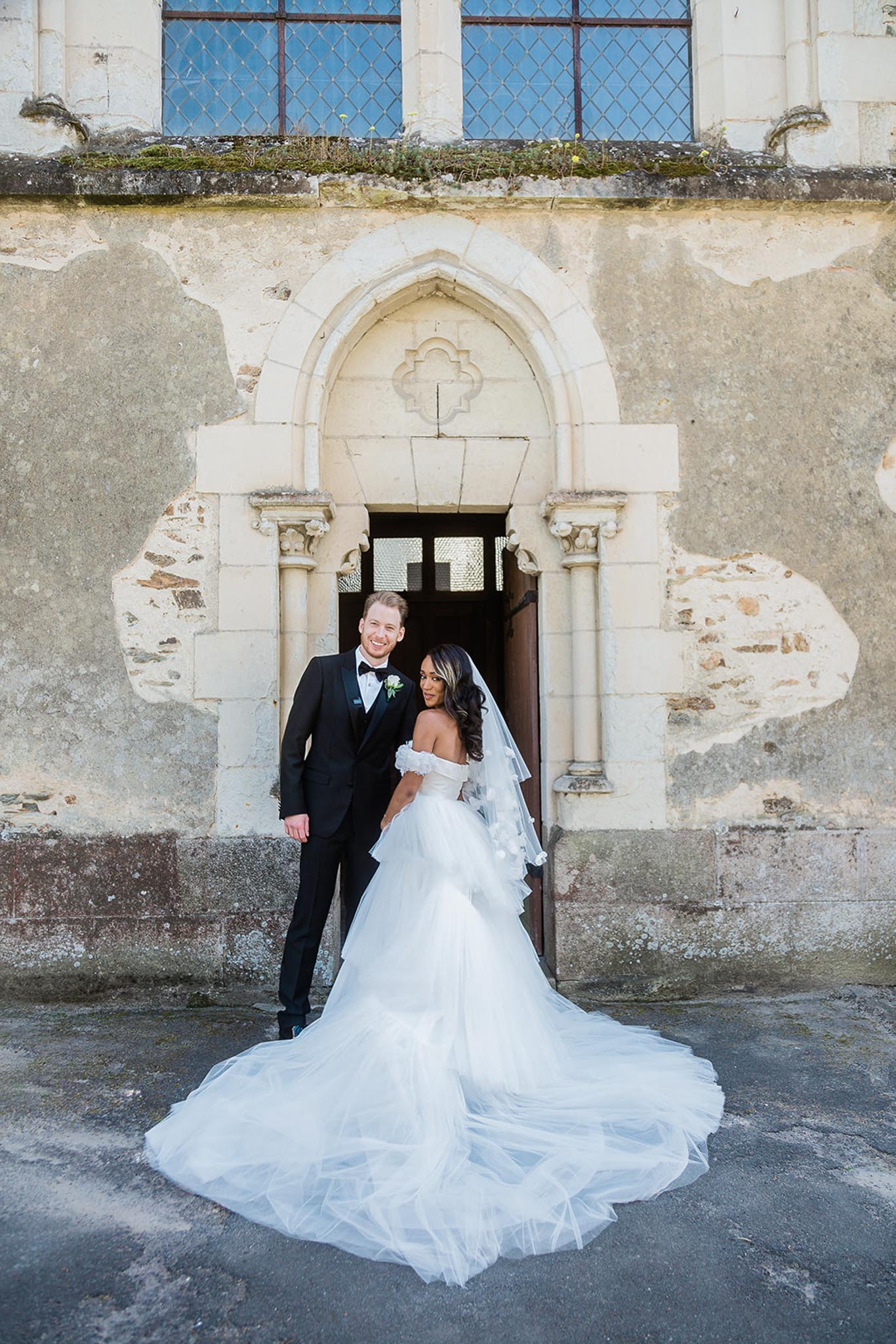 A couple portrait taken outdoors in front of a historic stone chapel or château chapel entrance featuring a Gothic-style pointed arch doorway with ornate carved detailing and flanking pilasters. The bride wears an off-the-shoulder white ballgown with a voluminous layered tulle skirt and an extended cathedral-length train spread across the ground, paired with a two-tier elbow-length veil and long dark wavy hair. The groom wears a classic black tuxedo with a black bow tie and a small white and green boutonniere. Both are facing the camera and smiling, standing close together in front of the arched doorway. The composition is a full-length portrait shot that emphasizes the dramatic spread of the bride's train in the foreground.