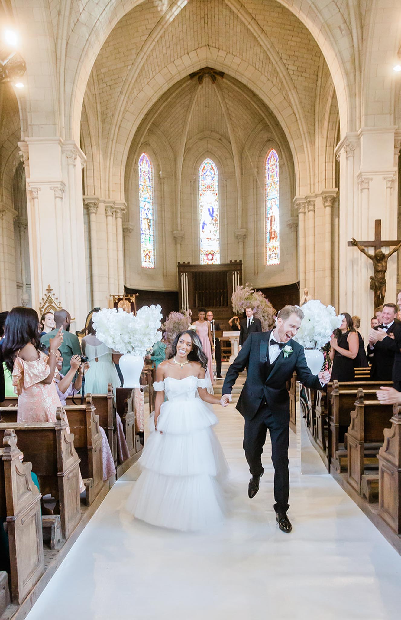 The bride and groom are walking back down the aisle in a joyful recessional inside a French Gothic stone chapel with vaulted arched ceilings, tall stained glass windows, and wooden pews. The bride wears an off-the-shoulder tiered white tulle ballgown and a gold statement necklace, while the groom wears a black tuxedo with a bow tie and a white floral boutonniere. Large arrangements of white hydrangeas are placed on white pedestals lining the aisle, and guests — dressed in a mix of blush sequin, sage green, and black formal attire — cheer and clap from the pews on both sides. The image is a wide shot taken from ground level looking toward the altar, capturing the full length of the aisle and the church's stone architecture.
