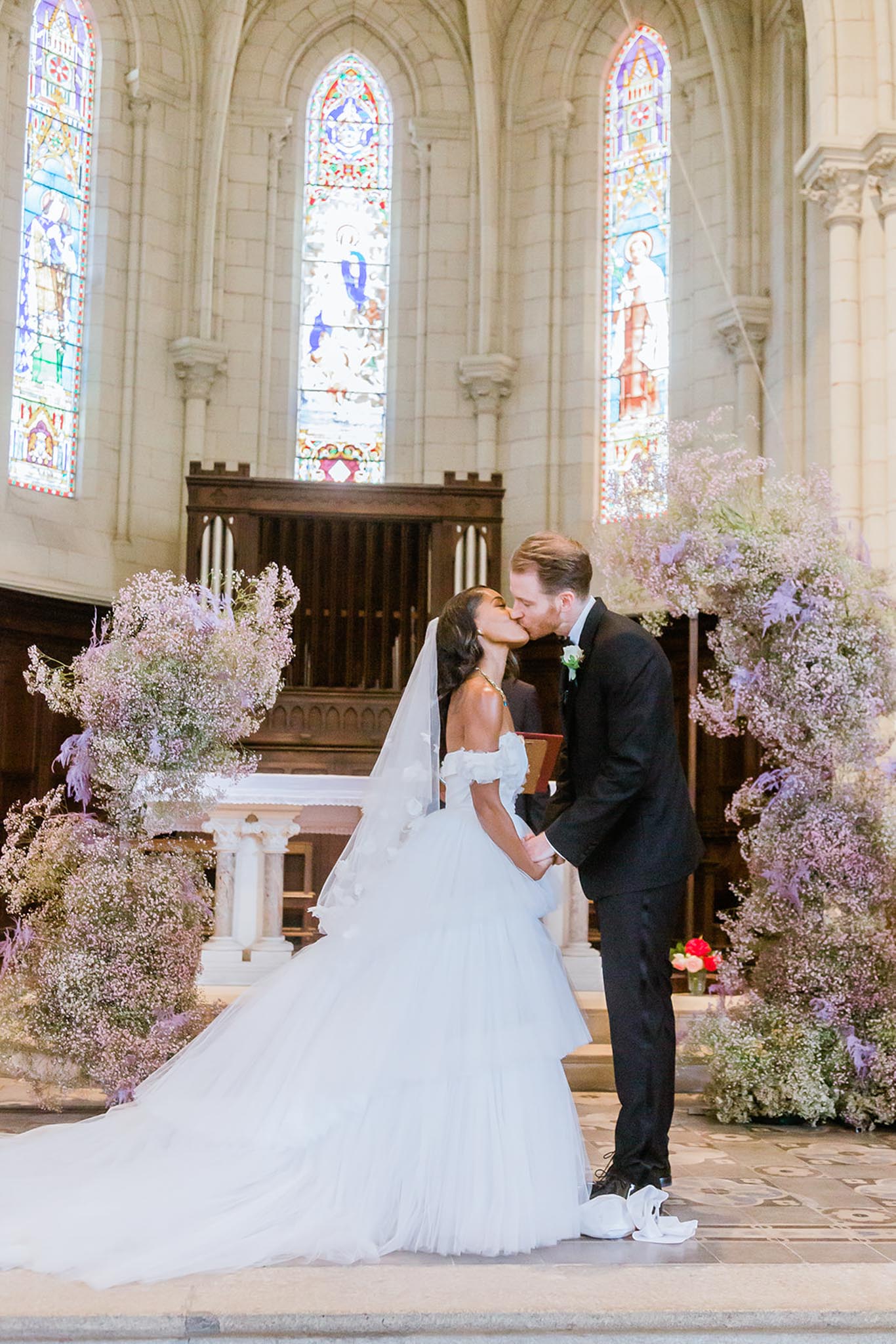 The bride and groom share their first kiss at the altar of a Gothic-style chapel with tall arched stained glass windows and stone architecture. The bride wears an off-the-shoulder white ball gown with a full tulle skirt, a cathedral-length veil, and a statement necklace, while the groom is dressed in a black tuxedo with a bow tie and white sneakers. Large asymmetric floral installations in soft lavender and white — appearing to be gypsophila, dried pampas-style grasses, and light purple blooms — flank both sides of the altar, creating a cloud-like, romantic ceremony backdrop. The composition is a mid-length portrait shot centered on the couple at the altar steps.