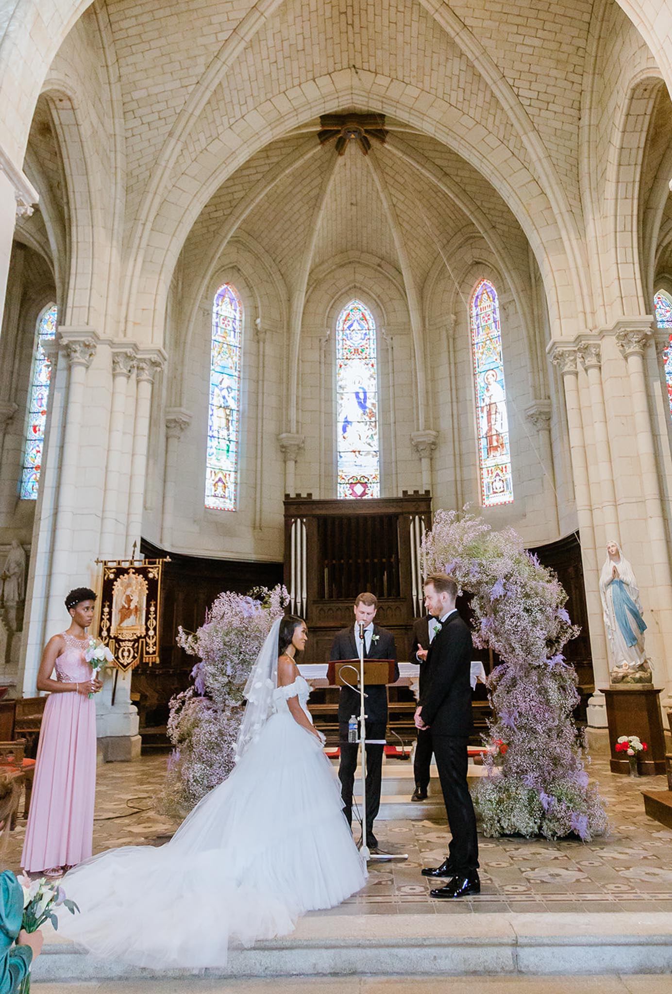 A wedding ceremony taking place inside a French Gothic stone church with ribbed vaulted ceilings, pointed arches, and colorful stained glass windows. The bride, wearing an off-the-shoulder white ballgown with a long veil and cathedral-length train, faces the groom, who is dressed in a black tuxedo, as an officiant speaks at a wooden lectern between them. Two large freestanding floral installations frame the altar, composed of cascading lavender and light purple limonium or dried-style blooms creating an arch-like effect on either side. A bridesmaid in a blush pink floor-length dress holds a small white bouquet to the left, and a partially visible guest in teal is seen at the lower left corner. The shot is a wide portrait-orientation image taken from the nave looking toward the altar, capturing the full architectural height and ceremony setup.