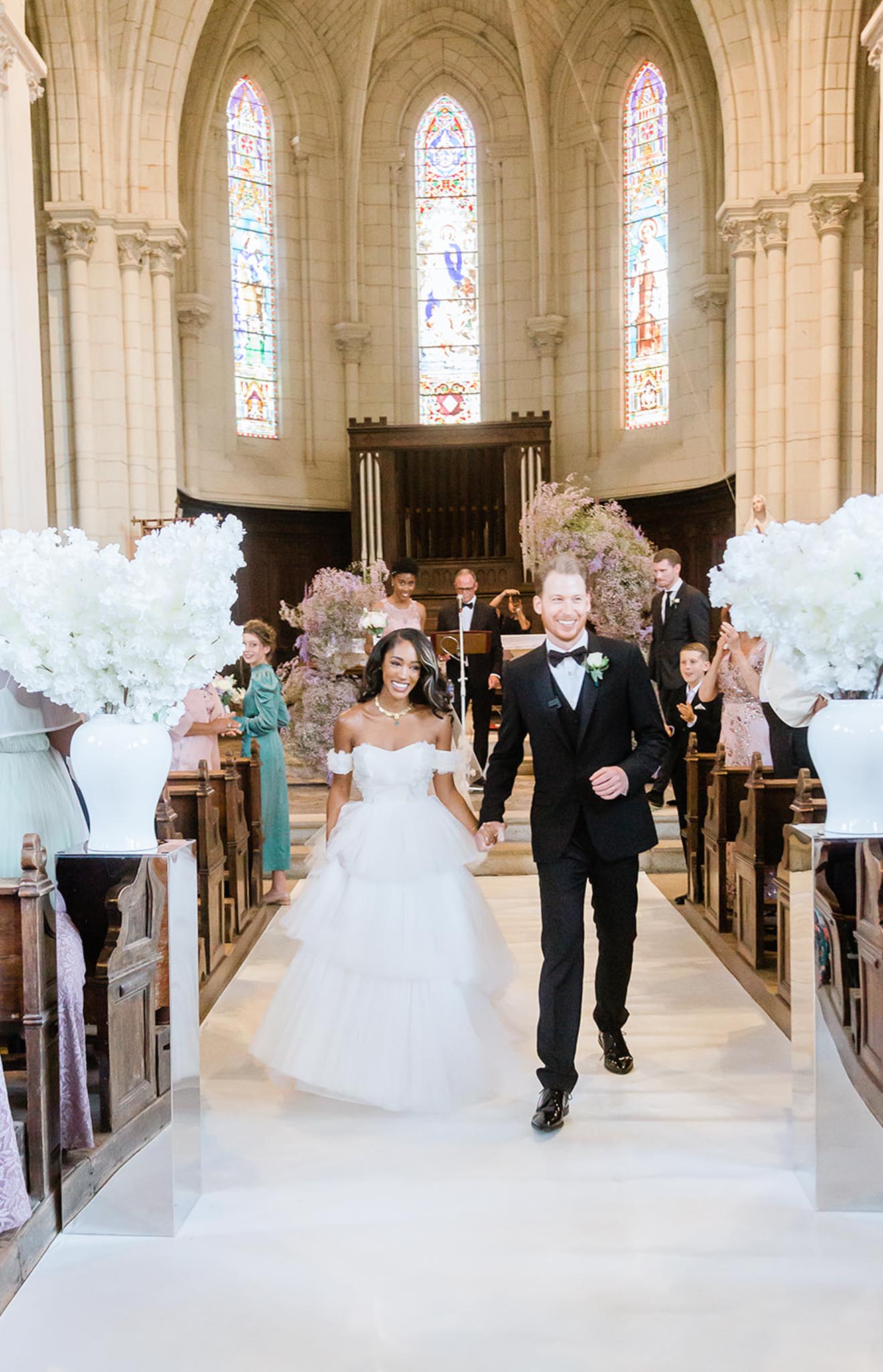 The bride and groom walk hand-in-hand down the aisle following their ceremony recessional inside a Gothic-style stone chapel with arched vaulted ceilings and colorful stained glass windows. The bride wears an off-the-shoulder white tulle ballgown with tiered layers and a gold necklace, while the groom wears a black tuxedo with a bow tie and a white boutonniere. Aisle pedestals on both sides hold large white arrangements of hydrangeas in white ceramic urns, and the altar in the background is decorated with tall arrangements of lavender and purple wisteria-style florals. Guests seated in wooden pews on either side are smiling and clapping, with a musician visible at a stand near the altar, captured in a wide portrait-orientation shot from a low central angle down the white aisle runner.