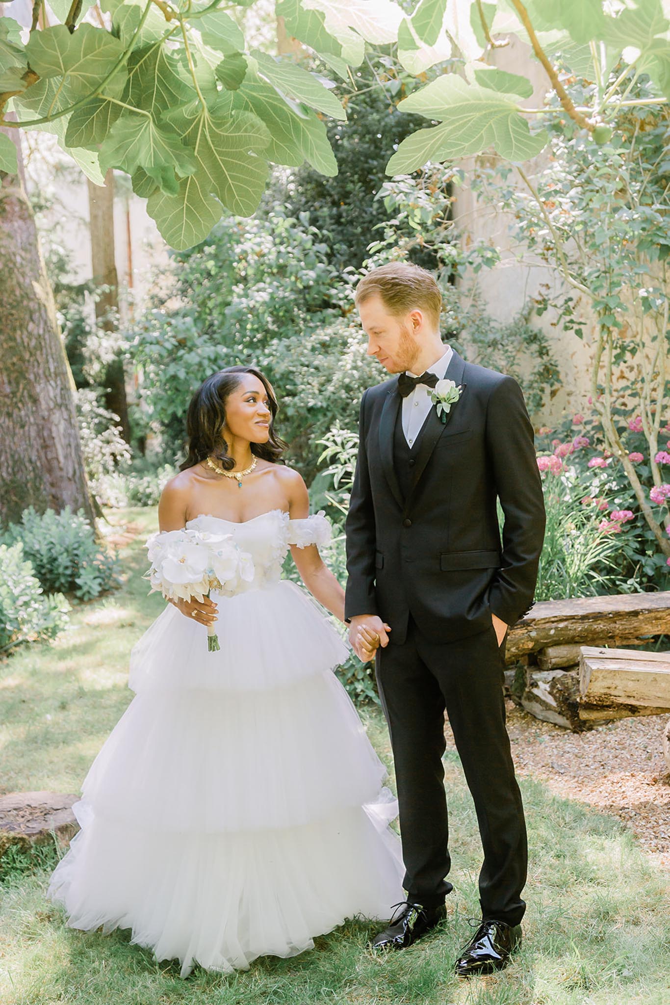 A couple portrait taken outdoors in a garden setting, with the bride and groom holding hands and looking at each other. The bride wears an off-the-shoulder white tulle ball gown with floral ruffle sleeve details, a gold and teal statement necklace, and carries a bouquet of large white peonies. The groom wears a black three-piece suit with a black bow tie and a white boutonniere with greenery. The shot is a mid-length portrait framed by large fig tree leaves overhead, with a stone wall and lush garden planting visible in the background.