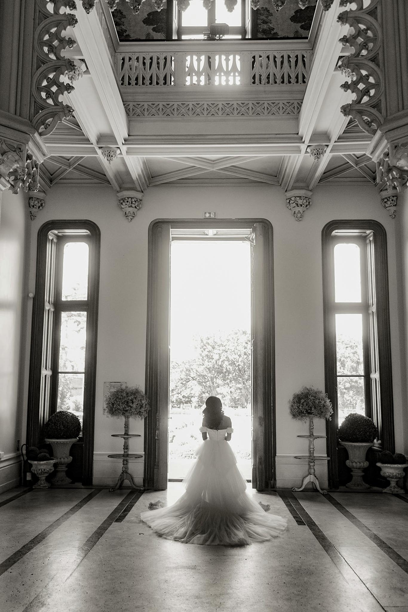 This black-and-white bridal portrait shows a bride standing alone at the center of a grand interior hallway, facing away from the camera toward a set of tall open double doors flooded with bright backlight. She wears an off-the-shoulder ball gown with a voluminous tulle skirt and an extended cathedral-length train that fans out across the tiled floor. The interior features ornate architectural detailing including decorative carved corbels, intricate latticed ceiling beams, a central skylight, and tall arched windows flanking the doorway. Potted topiary arrangements and floral arrangements on tall pedestal stands are placed symmetrically on either side of the entrance. The composition is a wide full-length rear portrait with strong contrast between the bright backlit doorway and the darker interior tones. Potential venue feature image.