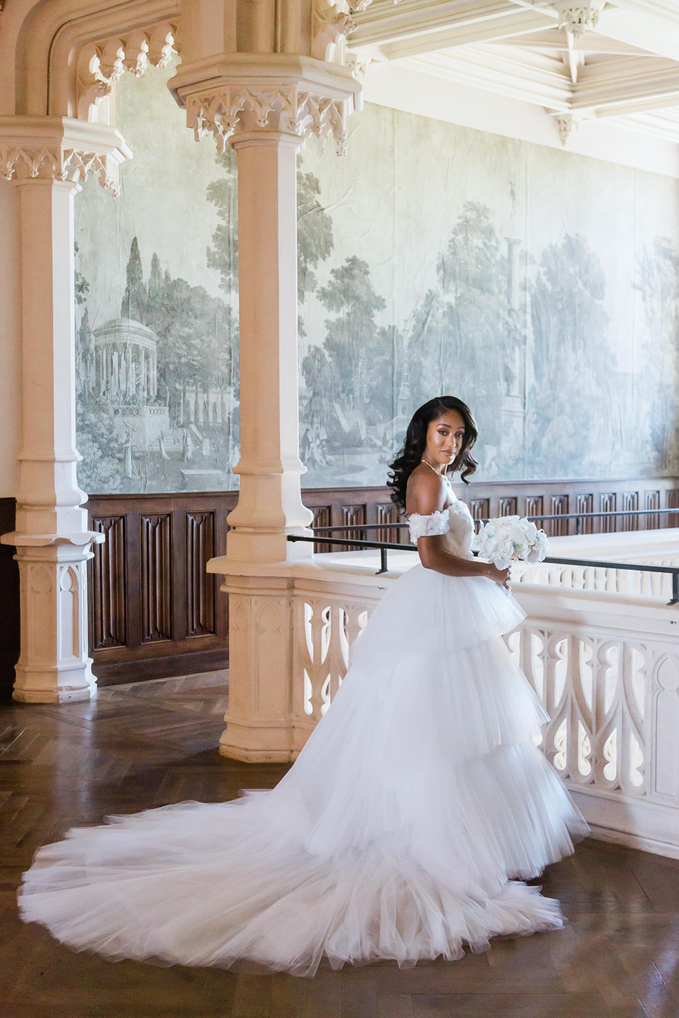 A bridal portrait taken indoors in what appears to be a historic chateau or castle interior, featuring an upper gallery or mezzanine level with ornate white Gothic-style carved stone columns and balustrade, dark wood paneling, herringbone parquet flooring, and large-scale grisaille landscape murals painted directly on the walls in muted blue-grey tones. The bride stands three-quarter length facing away from camera and looking back over her shoulder, wearing a white off-the-shoulder tiered tulle ballgown with a long cathedral train spread across the floor, accessorized with a pearl necklace, and holding a compact white bouquet of roses and soft white blooms. The composition is a full-length portrait shot with natural light filling the space from the right. Potential venue feature image.