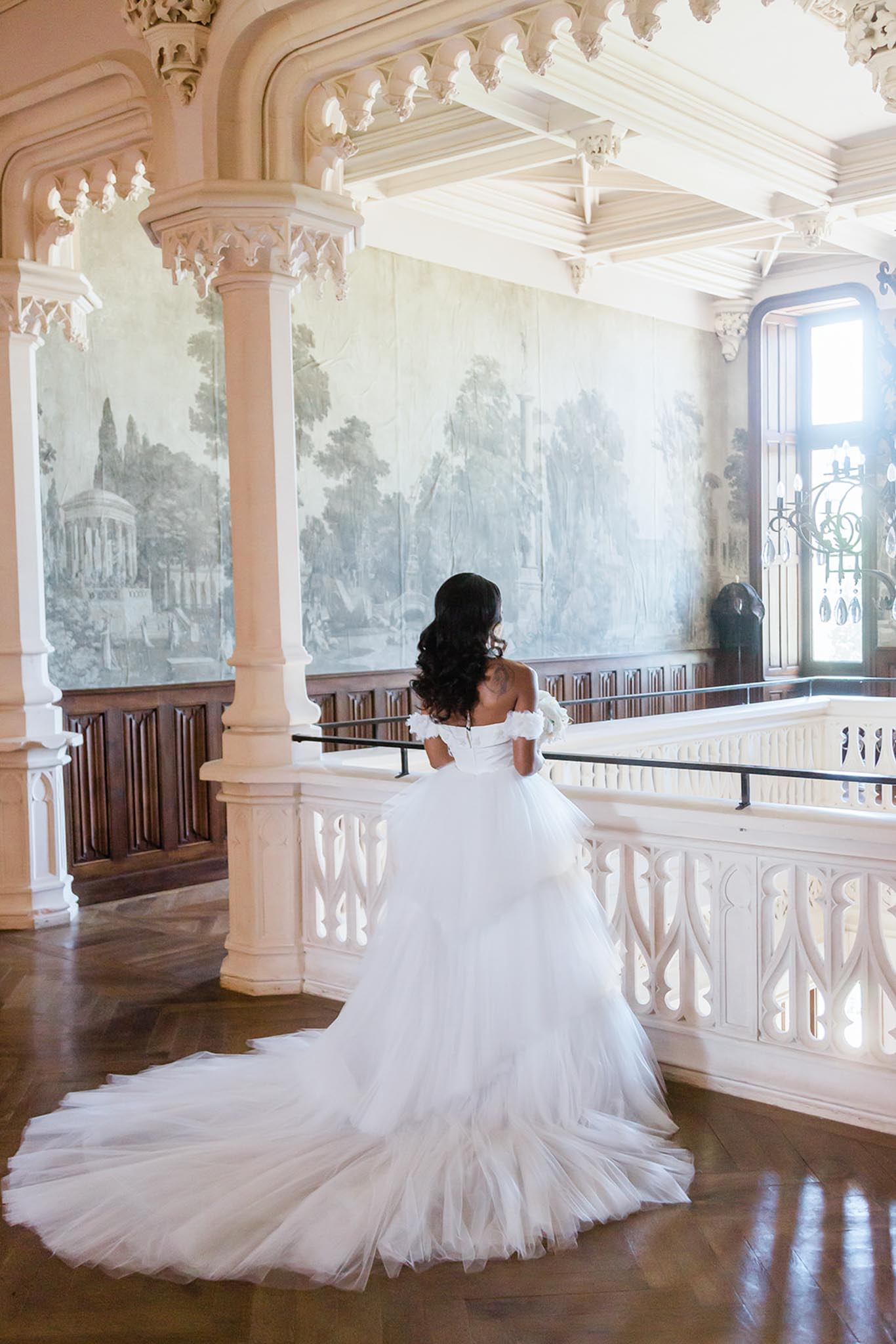 A bridal portrait taken indoors at what appears to be a historic chateau or palace, featuring an upper-level gallery or mezzanine with ornate white plaster columns, carved Gothic-inspired balustrades, and a herringbone parquet wood floor. The bride stands with her back to the camera, facing a large-scale grisaille landscape mural painted in muted blue-grey and green tones depicting classical architectural scenes. She wears a white off-the-shoulder ball gown with layered tiered tulle skirt and an extended cathedral train spread across the floor, with floral detailing at the sleeves; her long dark hair falls in waves down her back. She holds a small white bouquet and leans gently against the balustrade, with natural light entering through an arched window to the right. The composition is a full-length portrait shot from behind, emphasizing the volume of the gown against the ornate interior architecture. Potential venue feature image.