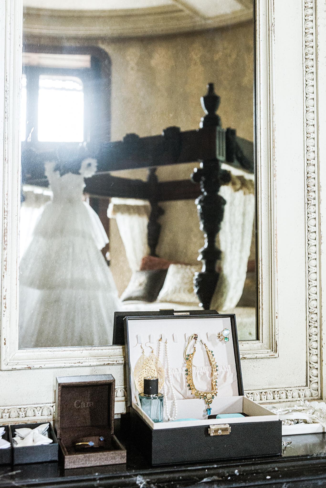 A getting-ready detail shot taken indoors, showing a dark dressing surface arranged with bridal accessories in front of a large ornate cream-painted mirror with carved detailing. In the foreground, an open black jewelry box displays gold statement earrings, a gold and emerald-toned necklace, pearl strands, and a floral gold accessory, alongside a small perfume bottle and a brown ring box labeled 'Cara' containing a ring with a blue stone. The mirror's reflection reveals a full white ballgown-style wedding dress hanging in the room, as well as a dark wood four-poster bed with cream draping, suggesting a chateau or historic manor bedroom setting. The composition is a medium close-up styled flat-lay perspective emphasizing the intentional arrangement of bridal jewelry and accessories.