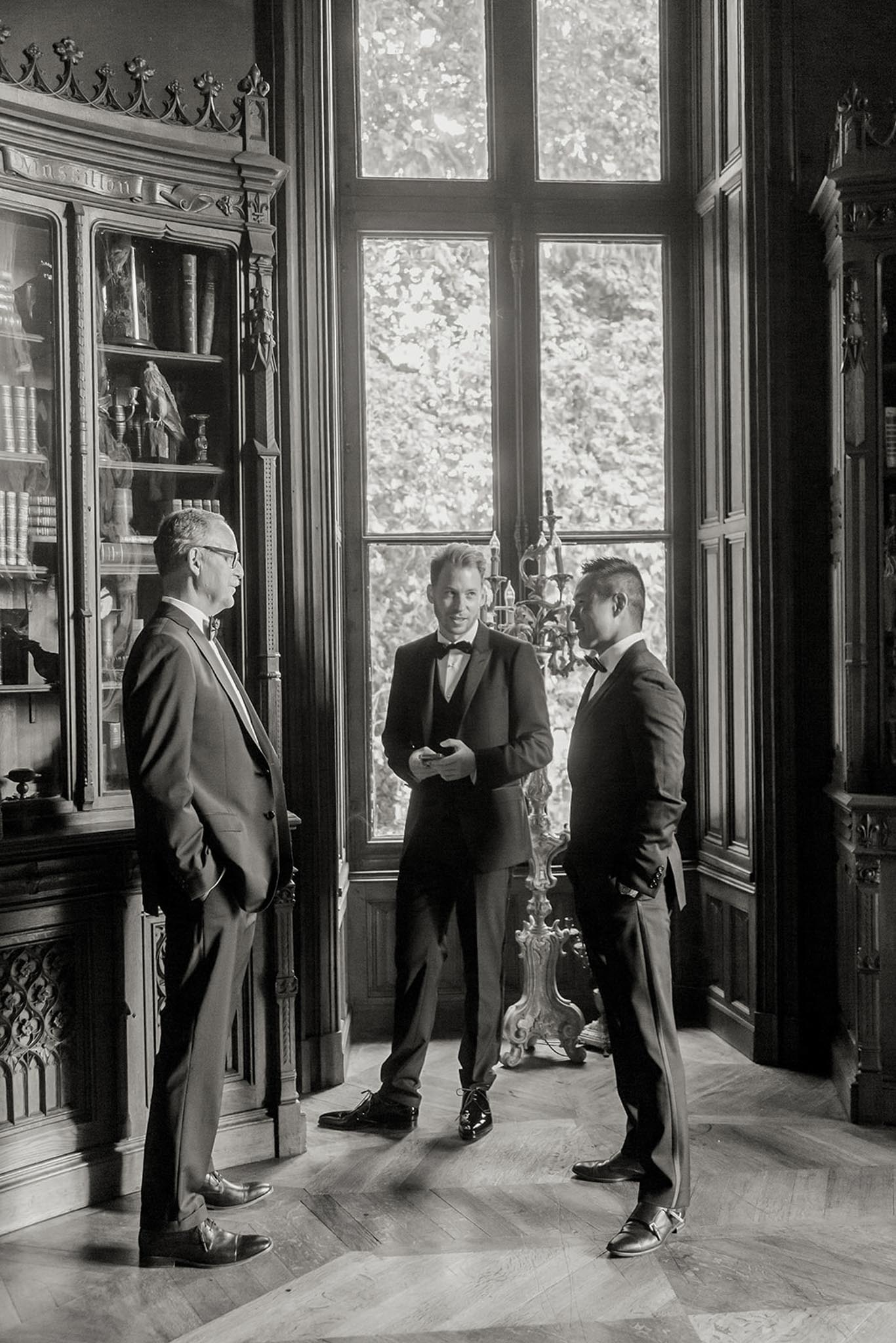 This black-and-white image shows three men in dark formal suits and bow ties conversing indoors, likely during a pre-ceremony getting-ready or cocktail moment. The setting is a grand chateau interior featuring ornate dark wood paneling, a tall arched window with multi-pane glazing, herringbone parquet flooring, an elaborate gothic-style bookcase filled with antique books and objects, and a decorative wrought-iron candelabra. The man in the center, who appears to be the groom, wears a three-piece suit and holds what looks like a phone, while the other two men — one older wearing glasses — stand facing him in conversation. The high-contrast tones emphasize the richly detailed architectural woodwork and the backlighting from the tall window. Portrait-style, full-length shot. Potential venue feature image.