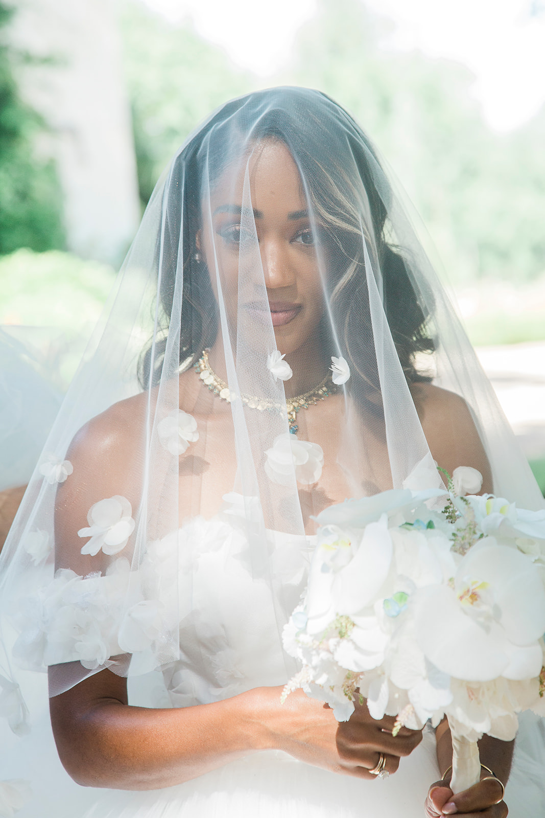A close-up bridal portrait taken outdoors with a softly blurred background. The bride is photographed under a sheer tulle veil that drapes over her face and body, adorned with small white 3D floral appliqués scattered across its surface. She wears an off-the-shoulder white gown with matching oversized floral appliqués and holds a bouquet of large white orchids and airy white blooms with subtle pale green accents. A gold and gemstone statement necklace is visible at her collarbone, and she wears gold rings on her fingers. Her hair is worn down in loose waves with a light blonde highlight framing her face. The overall styling is classic and refined, with an all-white palette throughout the dress, veil, and bouquet.