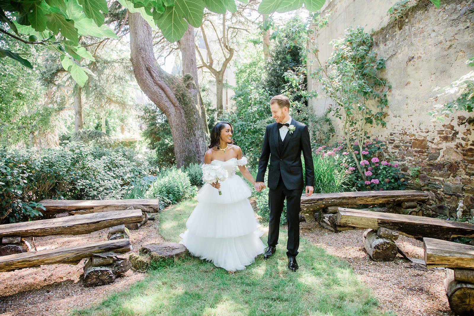 A couple portrait taken outdoors in a walled garden setting with rustic log benches lining a gravel path. The bride wears an off-the-shoulder white tiered tulle ballgown and carries a bouquet of white roses and ivory blooms, accessorized with a gold necklace. The groom wears a black tuxedo with a bow tie and a white boutonnière. The two are walking hand in hand, smiling at each other. The setting features aged stone walls with climbing vines, pink flowering shrubs, and mature trees framing the scene, giving a rustic yet refined atmosphere. The image is a medium full-length portrait shot in natural daylight.