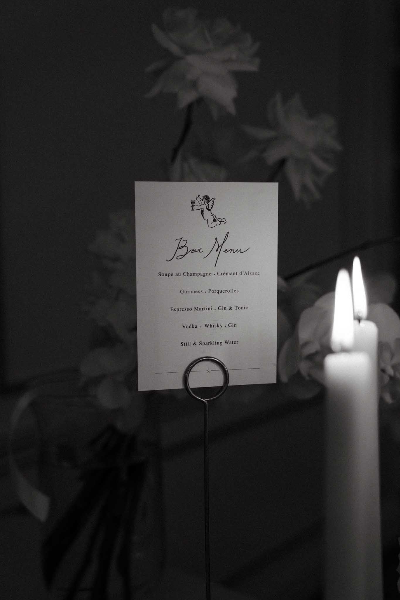 A black-and-white close-up detail shot of a wedding bar menu card displayed in a wire ring holder, positioned next to a lit pillar candle. The printed card features a cherub illustration at the top and lists drinks including Soupe au Champagne, Crémant d'Alsace, Guinness, Porquerolles, Espresso Martini, Gin & Tonic, Vodka, Whisky, Gin, and Still & Sparkling Water, written in a script and serif typeface combination. Soft floral arrangements are visible in the dark background, slightly out of focus. The image has deep shadow tones with bright highlights from the candle flame, giving it a moody, low-light atmosphere consistent with an indoor evening reception setting.