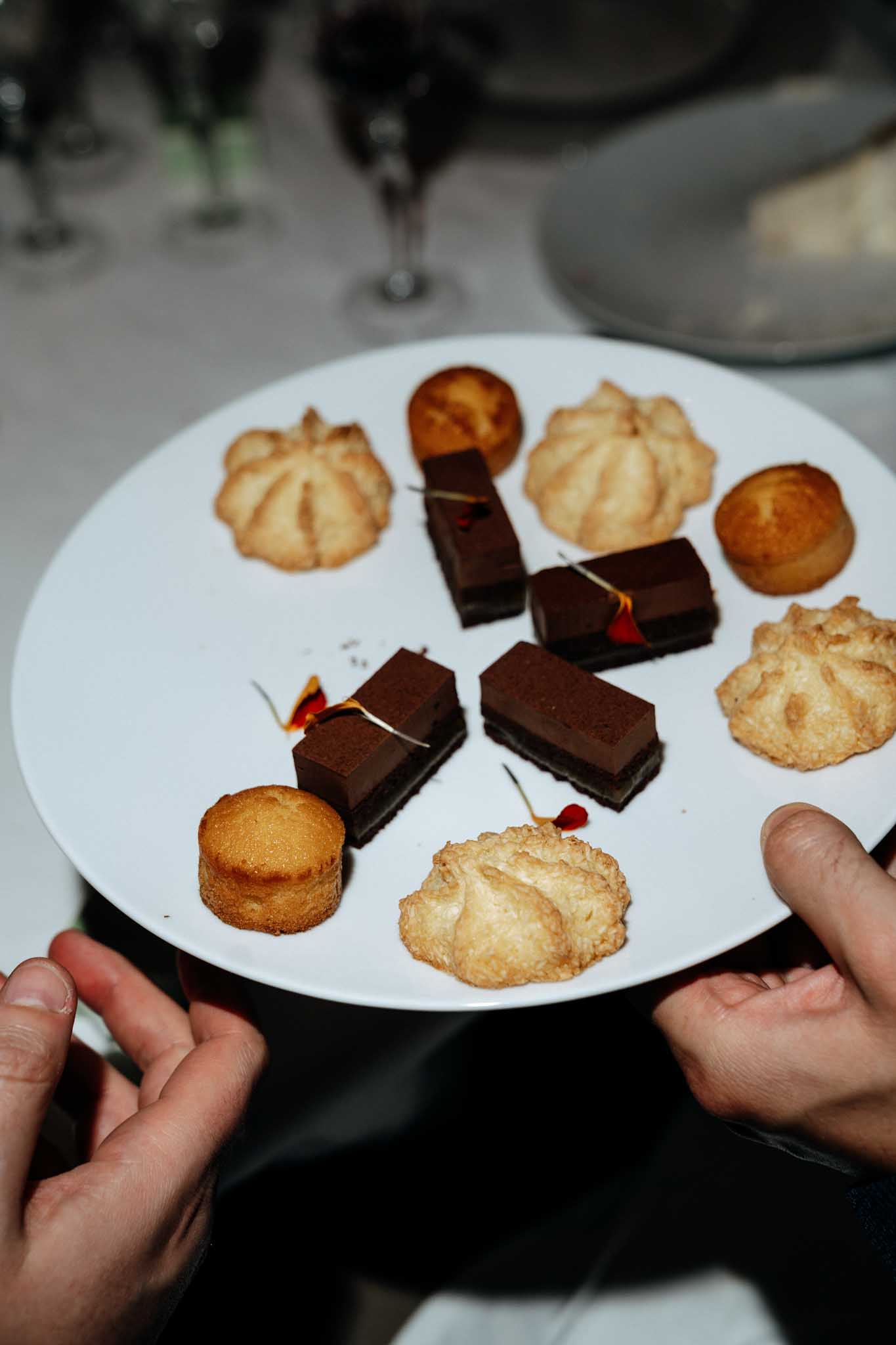 A close-up detail shot of a white round plate of mignardises being held by two hands during what appears to be a wedding reception dinner service. The plate holds an assortment of petit fours including dark chocolate ganache rectangles garnished with small red and orange dried flower petals on toothpicks, golden coconut macaroons, small round financiers or mini cakes, and cream puff-style choux pastries. Wine glasses and white table linens are visible in the soft-focus background, indicating an indoor reception setting with a formal table setup.