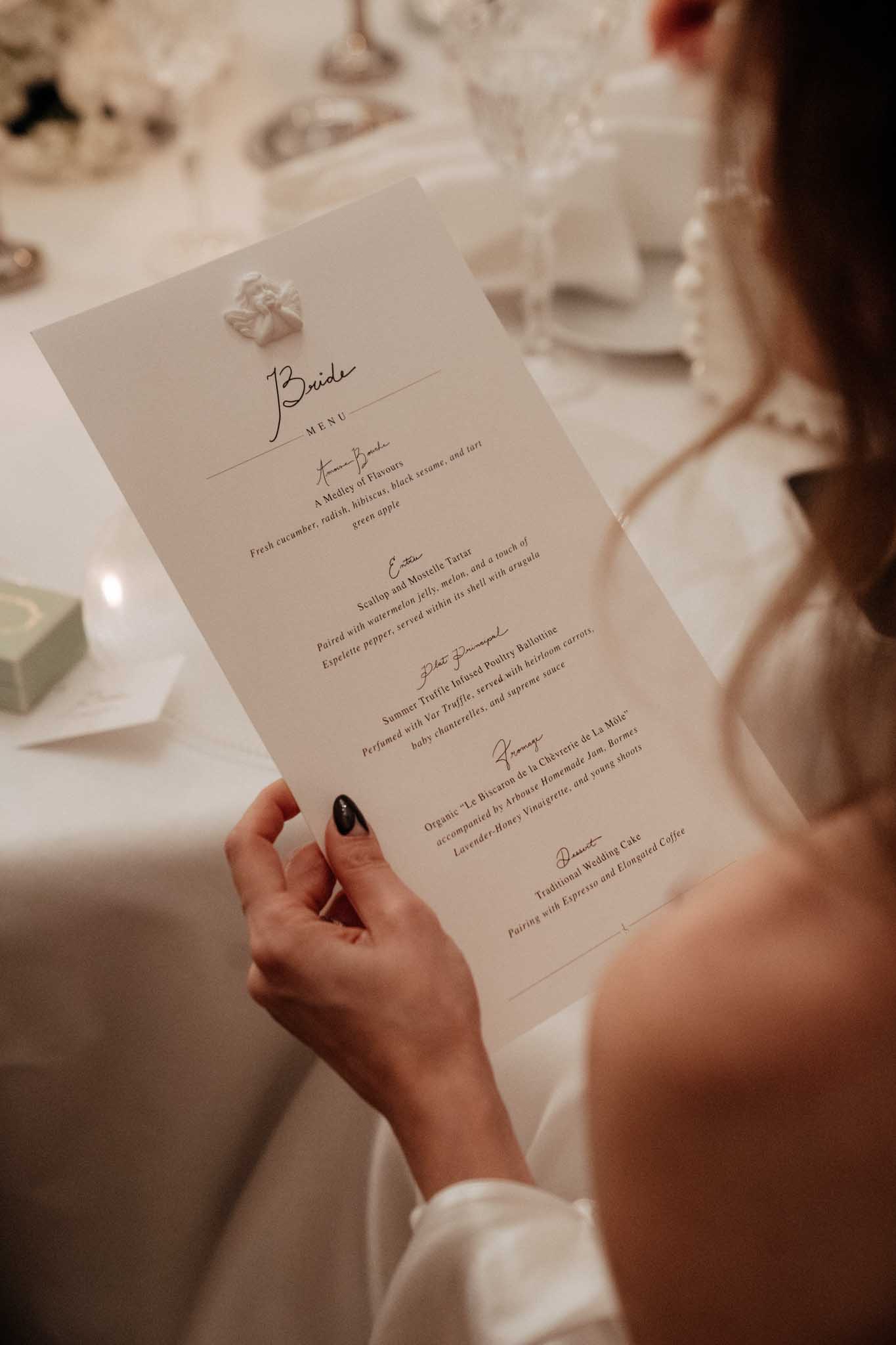 Close-up portrait shot of a bride reading her personalized wedding reception dinner menu, labeled 'Bride' at the top and decorated with a small sculptural white bow or floral embellishment. The menu is printed on cream card stock in a mix of script and serif typography, listing five courses including an amuse-bouche, entrée of scallop and mostelle tartare, a main of summer truffle infused poultry ballottine, a cheese course, and a dessert of traditional wedding cake. The bride wears a white satin gown with long sleeves and pearl drop earrings, with dark-painted nails visible as she holds the card. The reception table in the background features white linen, crystal glassware, candles, and white floral arrangements, suggesting a classic, formal indoor reception setting.