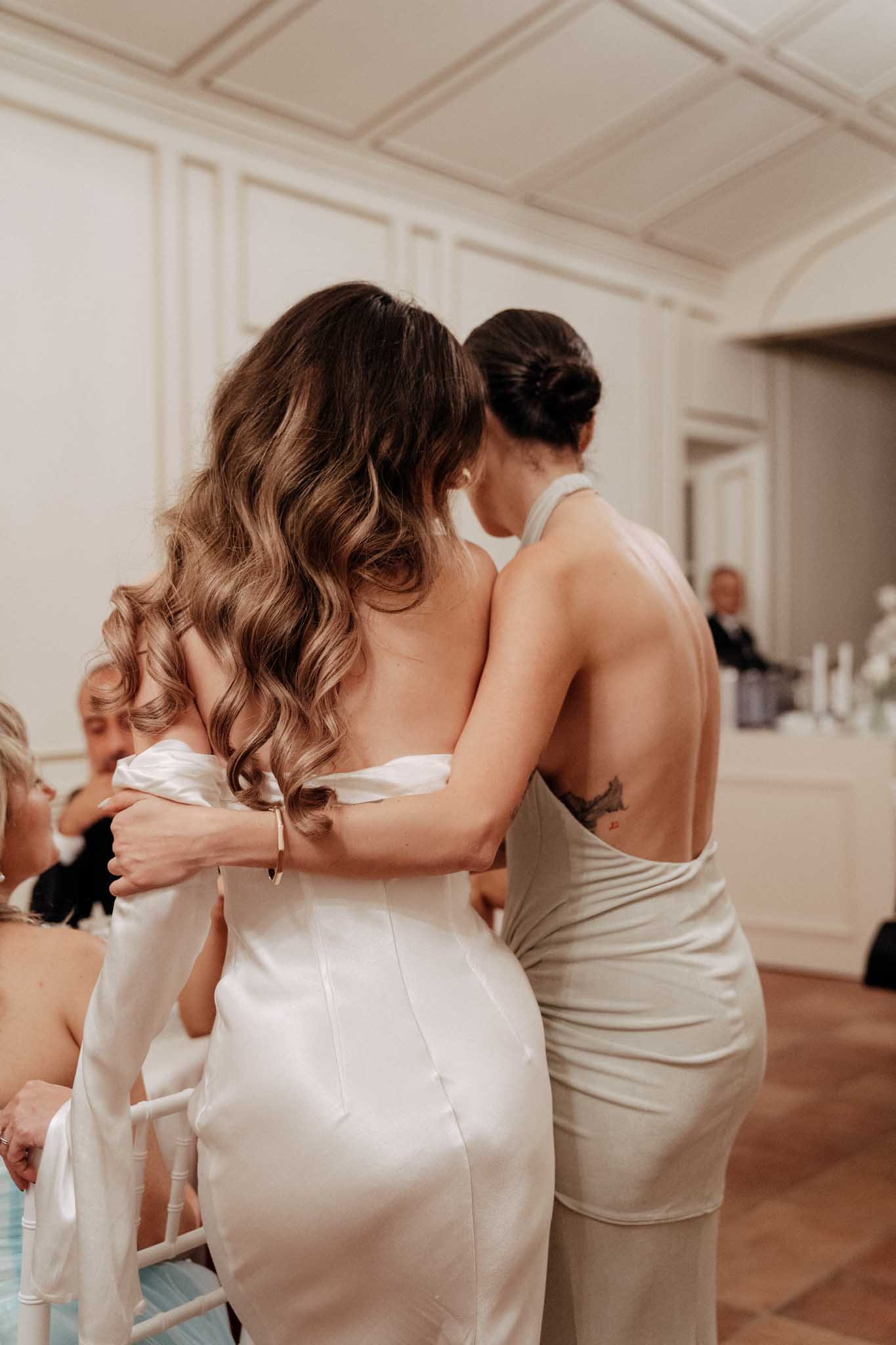 A candid reception moment captured from behind, showing a bride in a white satin strapless fitted gown being embraced by a woman in a backless halter-neck champagne/sage fitted dress with a visible tattoo on her back. The bride has long, loosely curled highlighted brunette hair worn down. Both women are facing away from the camera in what appears to be a warm embrace. The setting is an indoor ballroom or formal reception room with white paneled walls, coffered ceiling, and parquet wood flooring. Several seated guests are partially visible in the background, along with a bar area. The overall styling is modern and minimal with a neutral ivory and champagne palette.