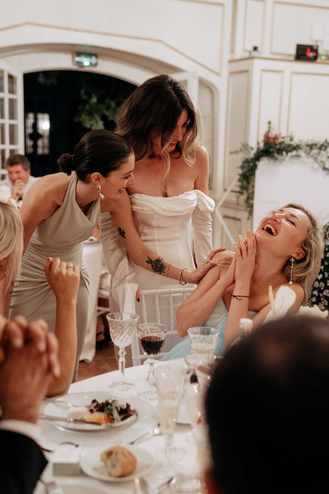 A candid reception moment captured indoors, likely in a chateau or manor ballroom with white paneled walls and an arched doorway visible in the background. The bride, wearing an off-the-shoulder white structured gown with a visible floral tattoo on her arm, leans over a seated guest who is laughing openly with her head thrown back; a bridesmaid in a silver-grey halter satin dress stands nearby smiling. The dinner table in the foreground is set with white linens, crystal glassware, red wine, and plated salad courses on silver chargers. The shot is a medium candid portrait capturing a genuine moment of laughter and connection among the women during the seated dinner reception.
