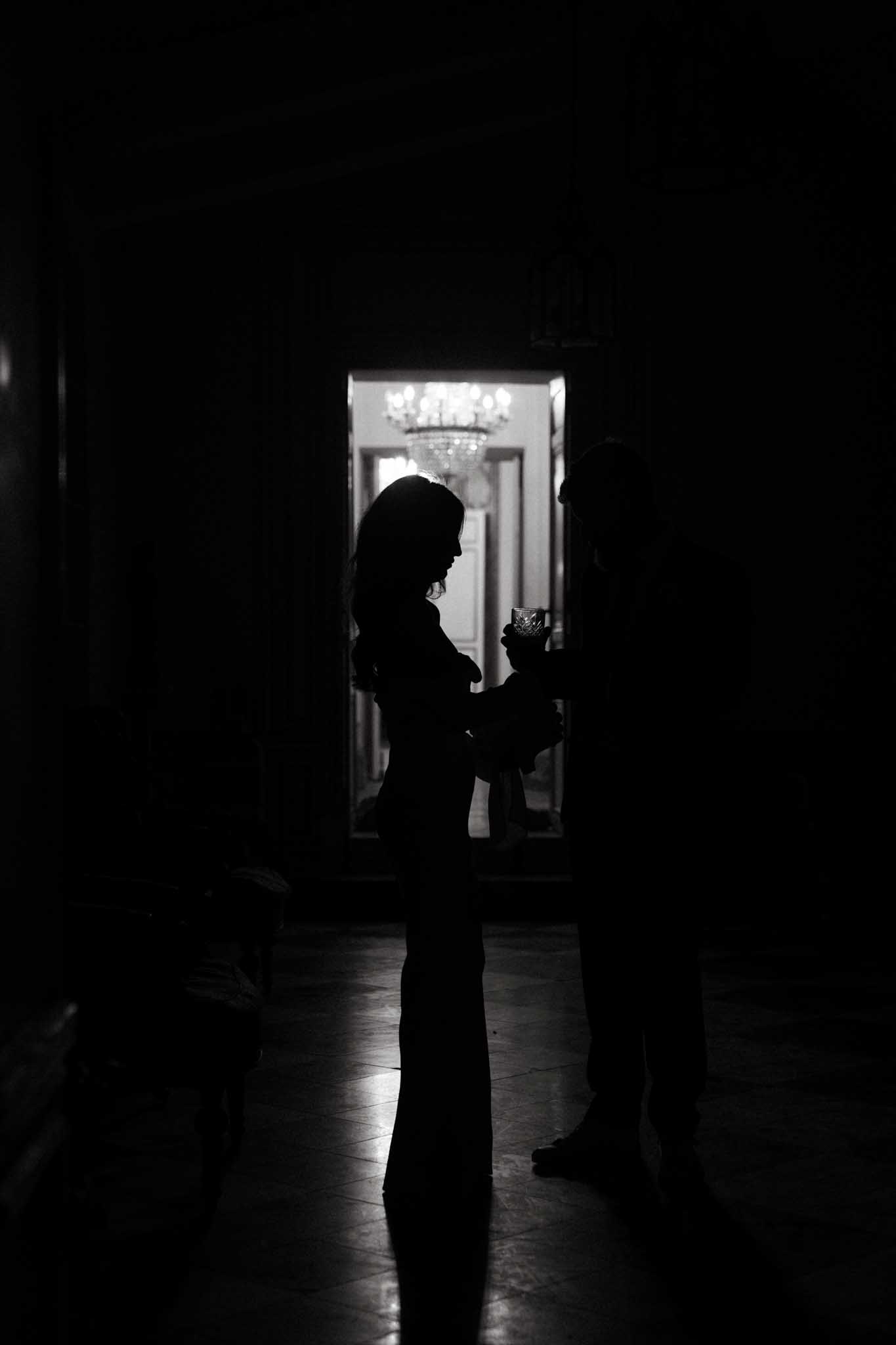 This black-and-white image shows two people silhouetted in a dark interior hallway or anteroom, one holding what appears to be a crystal glass. The figures are backlit by a brightly lit doorway behind them, through which a large crystal chandelier is visible, suggesting a formal chateau or grand mansion interior. The high contrast between the deep black foreground and the bright, warmly lit room beyond creates a dramatic tonal range. The wide shot captures the tiled floor reflecting a small amount of light, and a third person is partially visible crouching in the lower left corner.