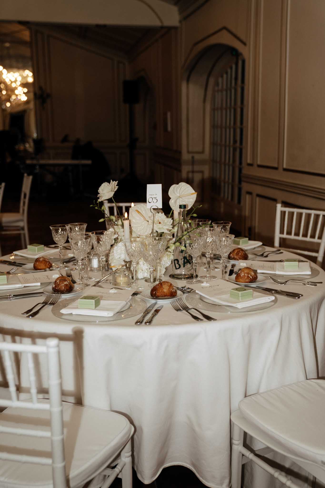 A close-up detail shot of a fully set wedding reception table, numbered table 03, inside a formal indoor venue with warm-toned wood paneling and arched architectural details visible in the background. The round table is covered with a white satin tablecloth and surrounded by white chiavari chairs with ivory cushions. Each place setting includes a silver charger plate, white linen napkin, crystal cut-glass stemware, and silver cutlery, with small mint green favor boxes and individual bread rolls placed at each seat. The centerpiece consists of white anthurium flowers, white roses, and white hydrangeas with trailing greenery, accompanied by two lit taper candles and a wine bottle used as a decorative element. The overall decor palette is white, ivory, and soft green with silver accents, reflecting a classic modern styling approach.
