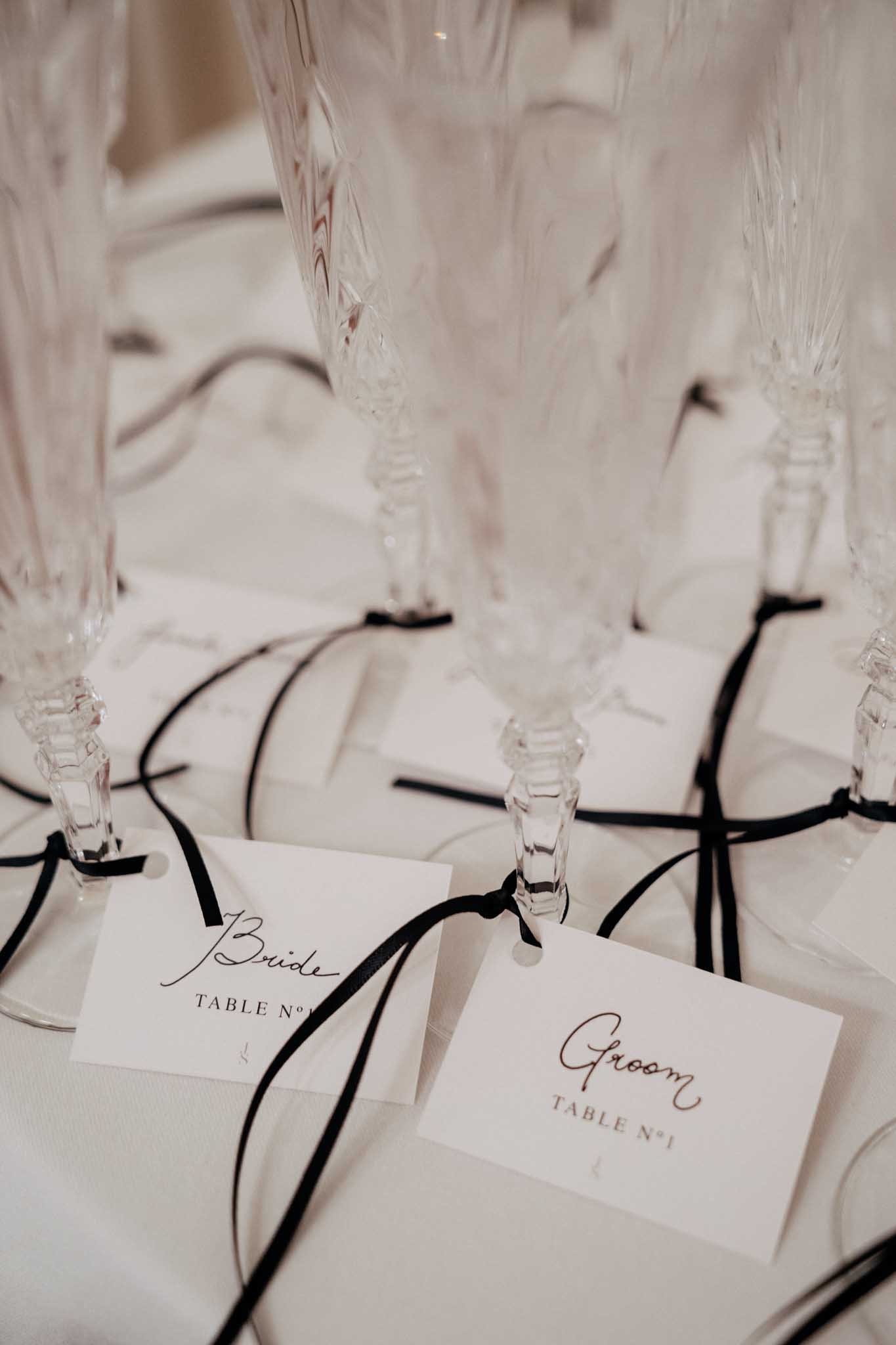 Close-up detail shot of a wedding reception table setting featuring crystal cut-glass champagne flutes with small white place cards tied to their stems using thin black satin ribbon. The two visible place cards read 'Bride, Table N°1' and 'Groom, Table N°1' in a mix of script and printed typography. The table is dressed in white linen, and the overall decor palette is monochrome — white and black — suggesting a classic, formal styling theme. The image is slightly shallow in focus, with the background flutes softly blurred.