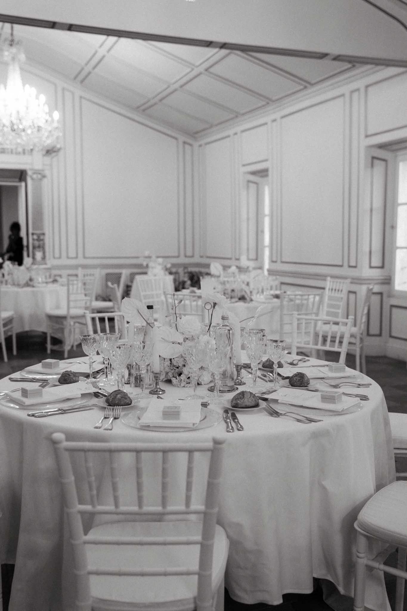 A black-and-white image of a wedding reception tablescape inside a formal ballroom with paneled white walls and a crystal chandelier visible in the upper left. The foreground shows a round table dressed in a floor-length white linen, set with white chiavari chairs, crystal cut-glass stemware, silver flatware, folded white napkins on charger plates, small favor boxes, and individual bread rolls at each place setting. The centerpiece features large white blooms — likely peonies or roses — with delicate branching stems and a glass bottle vase. Several additional round tables with identical styling are visible in the background, and a lone figure stands near the far wall. The overall decor palette is all-white with silver accents, consistent with a classic, formal French reception aesthetic. Wide shot from a slightly elevated angle. Potential venue feature image.