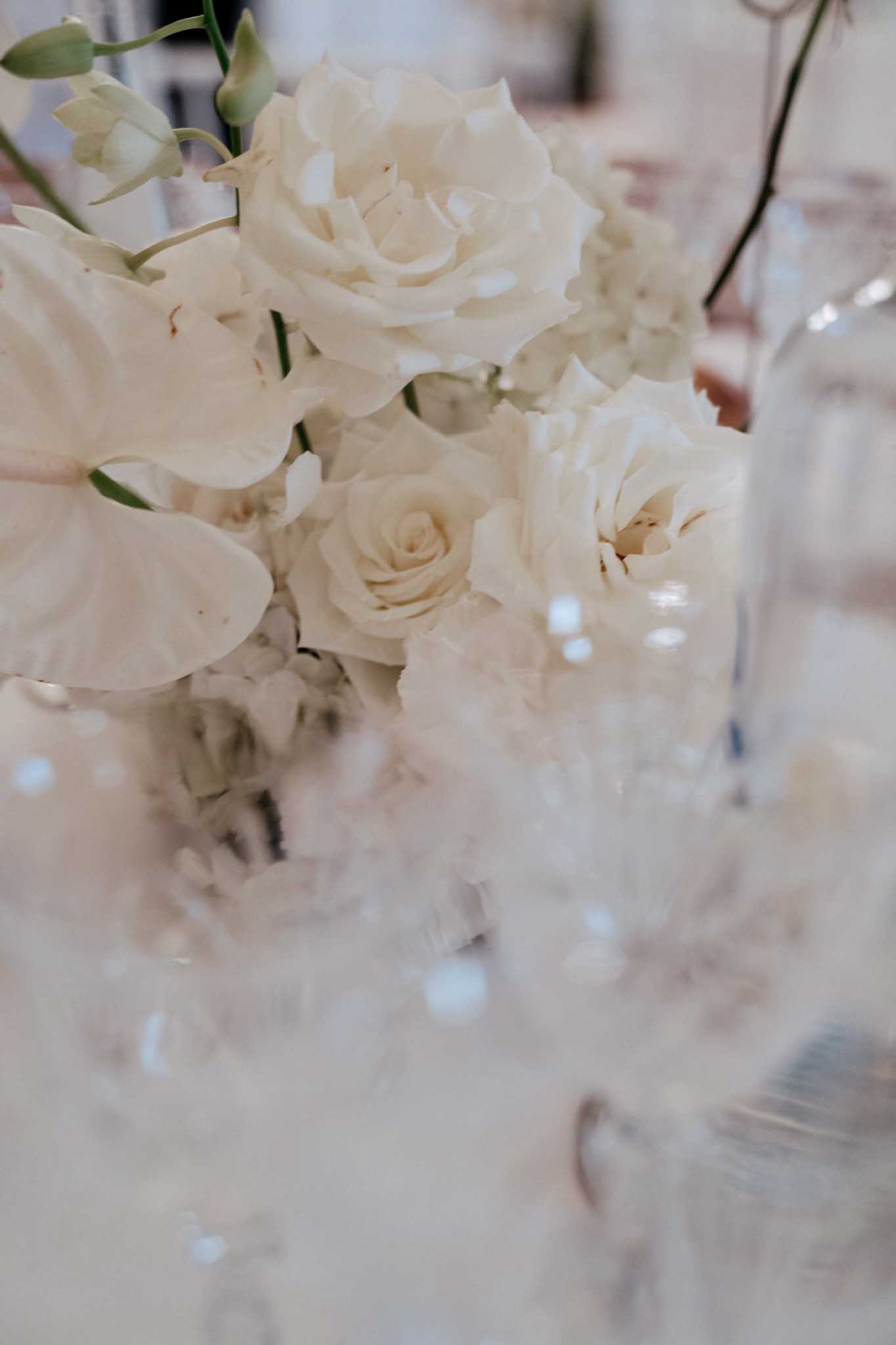 Close-up detail shot of a reception table centerpiece featuring cream garden roses and white phalaenopsis orchids with green stems, arranged in what appears to be a clear glass vase. The foreground is intentionally out of focus, revealing soft bokeh from glassware and crystal elements on the white linen-covered table. The overall decor palette is all-white and cream, consistent with a classic, minimal styling theme. Additional glassware and what appears to be a thin floral stem in a bud vase are visible in the blurred background.
