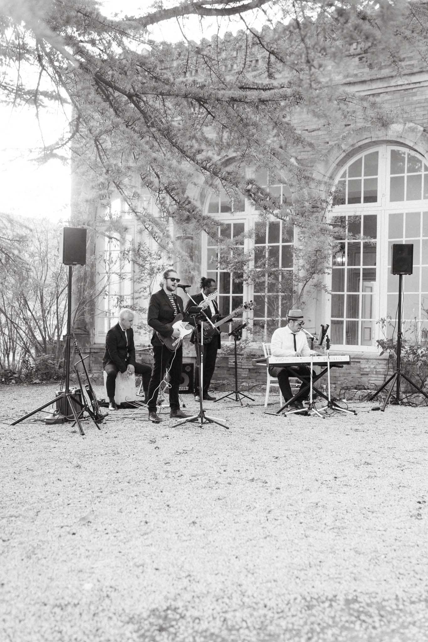 A black-and-white wide shot of a live band performing outdoors on a gravel courtyard during what appears to be a wedding cocktail hour or reception. Four musicians are visible: a guitarist singing at a microphone stand, a bass guitarist, a keyboard player seated at a portable keyboard wearing a hat and tie, and a percussionist seated behind a cajon. Two PA speakers on stands flank the group. The performance takes place in front of a stone chateau facade featuring large arched white-framed windows, with climbing plants visible on the building exterior. The image has bright, high-key tones with soft contrast.