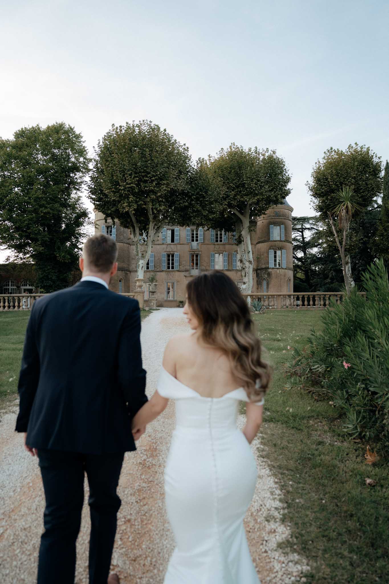 A couple portrait shot from behind as the bride and groom walk hand-in-hand along a gravel path toward a French chateau. The groom wears a dark navy suit and the bride wears an off-the-shoulder fitted white gown with a low open back and button detailing, her long wavy brunette hair loose down her back. The chateau is a multi-story stone building with blue shuttered windows, a rounded tower, and a balustraded terrace, framed by large plane trees. The couple are slightly out of focus in the foreground, with the chateau sharp in the background, creating depth in the composition. Potential venue feature image.